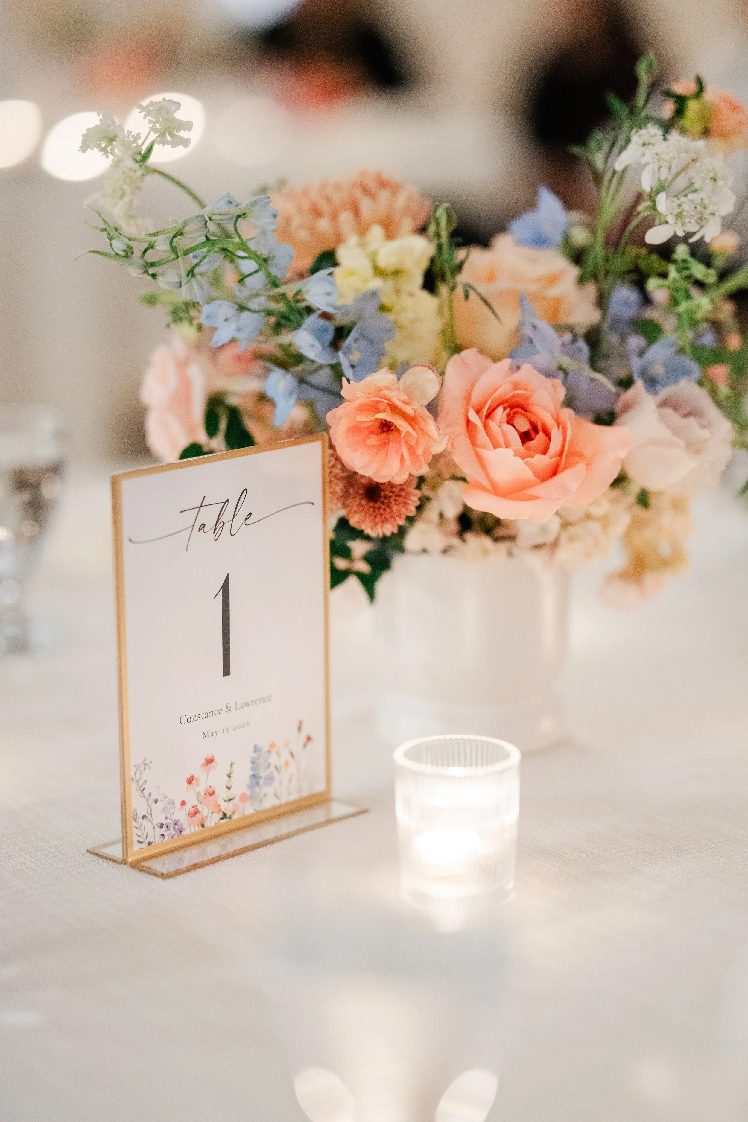 A floral centerpiece with pink, white, blue, and yellow flowers. A table number card with the number 1 and names, date, and floral illustrations. A small candle holder with a lit candle positioned in front of the card.