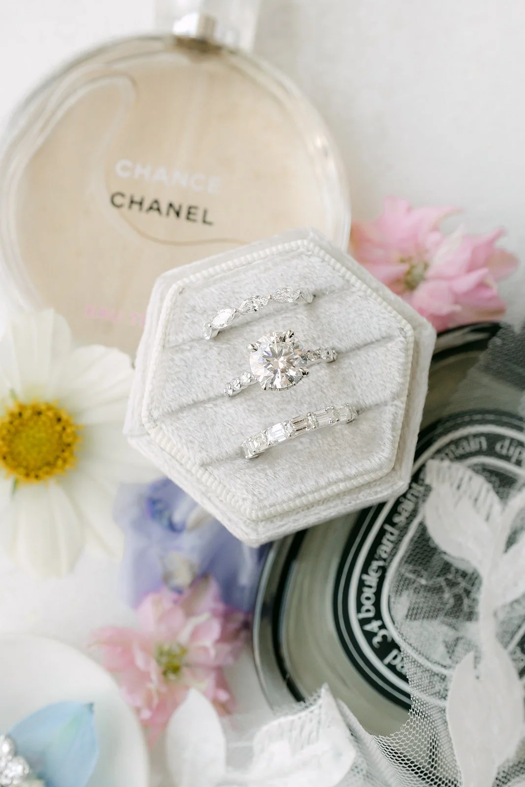 A hexagon ring box holding the wedding rings beside a bottle of perfume and pink petals