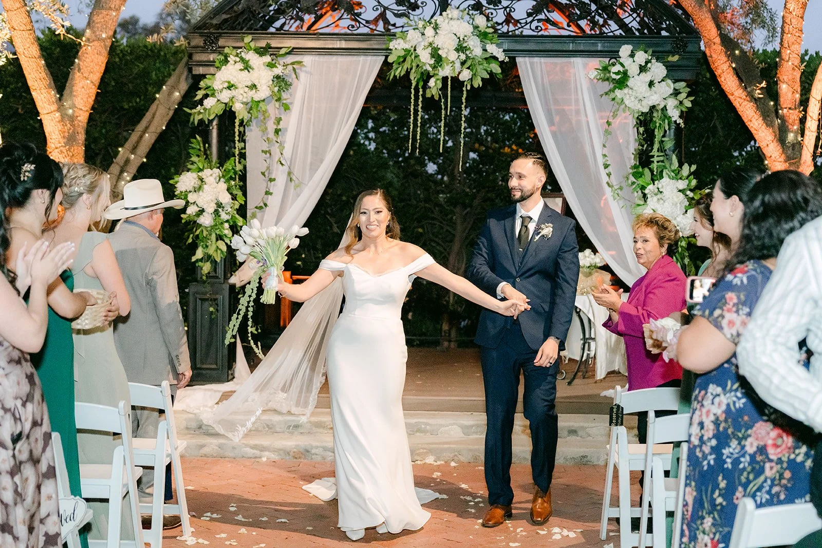 Mariana throwing her arms up as they walk back up the aisle, photographed by One and Only Production