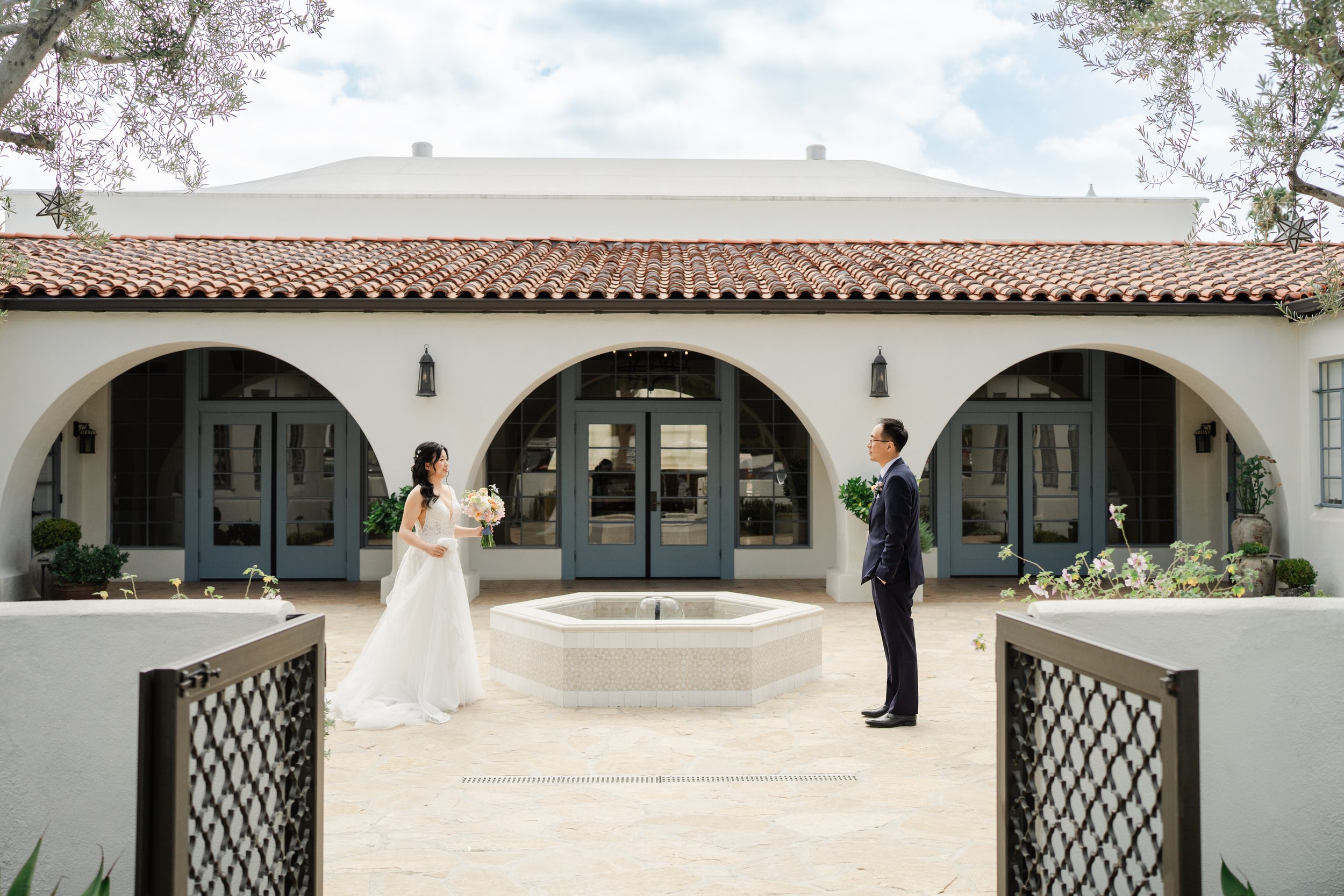 Bride and groom standing in front of a white building with arched windows and a tiled roof, holding flowers, during a wedding ceremony.