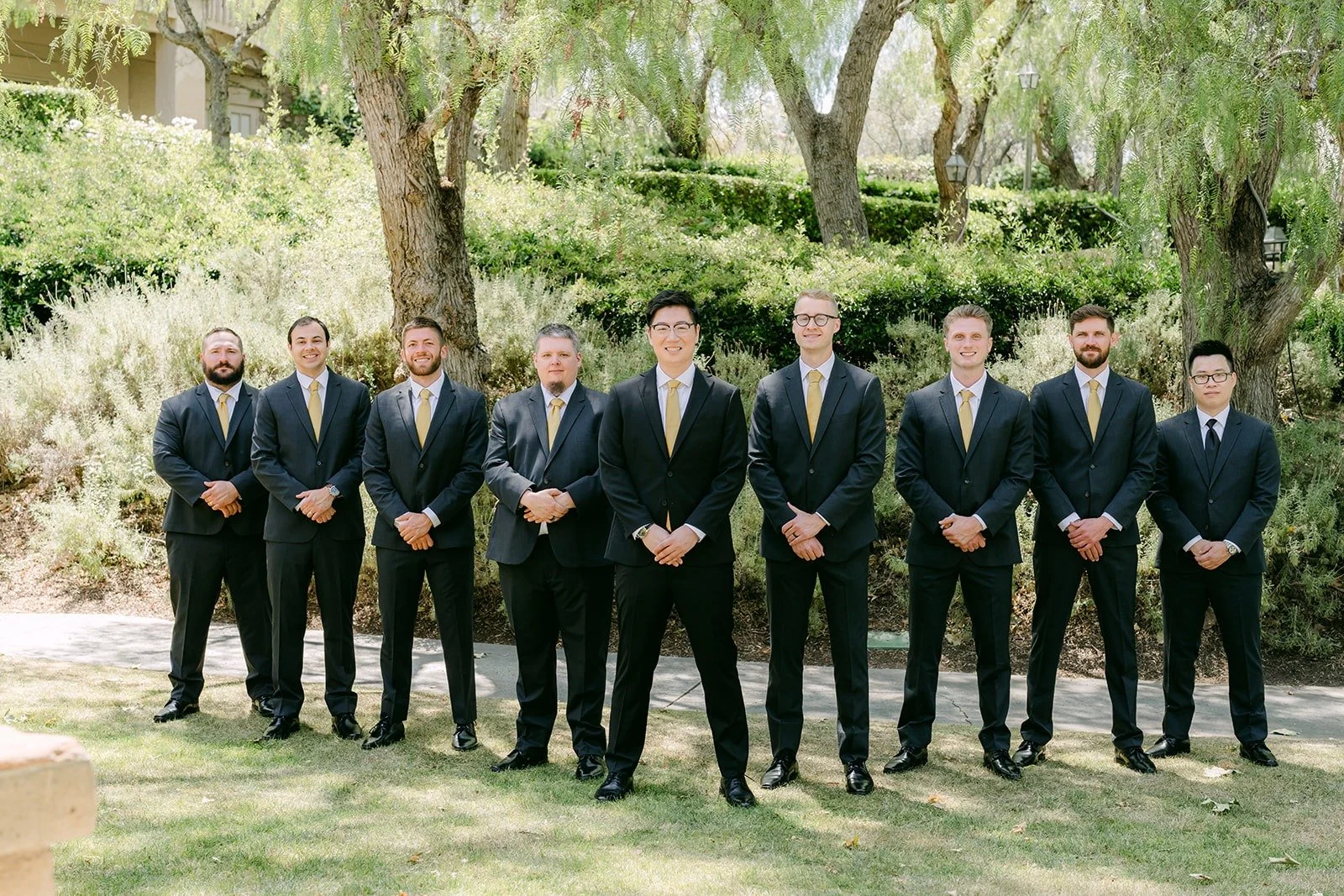 Zach and his groomsmen lined up in black suits and gold ties among the trees at Rancho Bernardo Inn