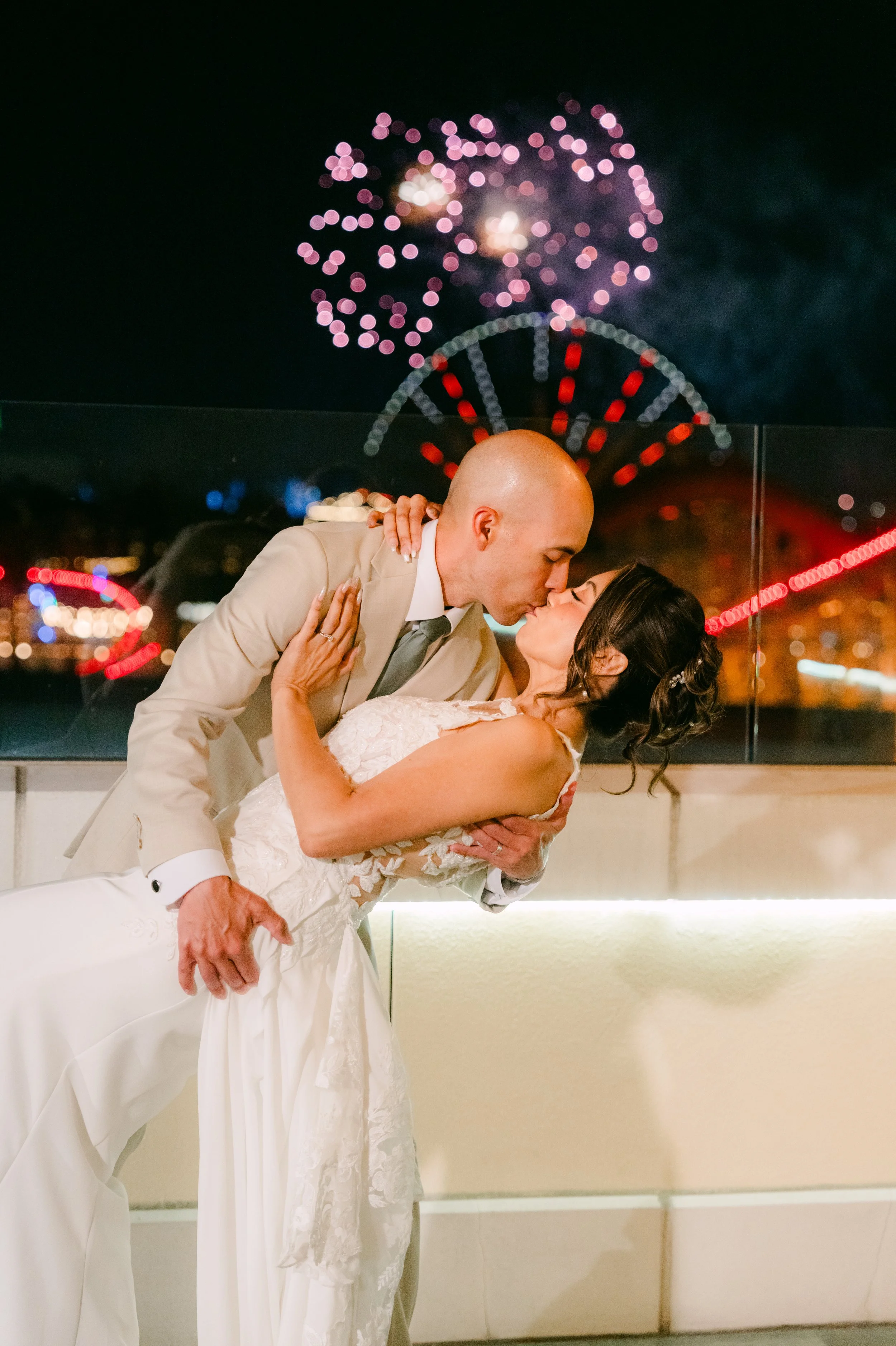 A newlywed couple sharing a kiss at night with fireworks in the background, the bride in a white wedding dress and the groom in a beige suit.