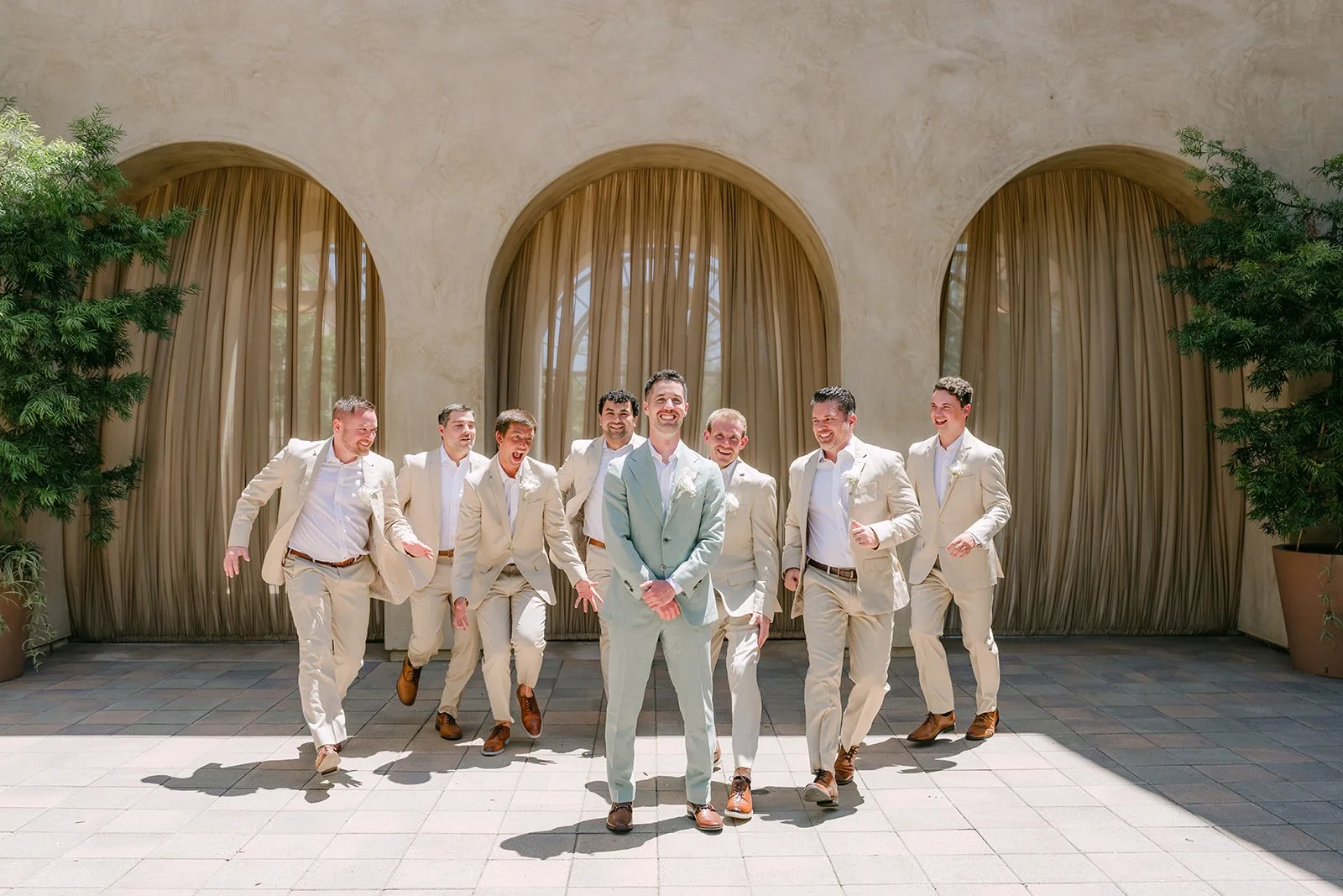 Groom and groomsmen in sand linen suits walking toward the camera past arched curtains