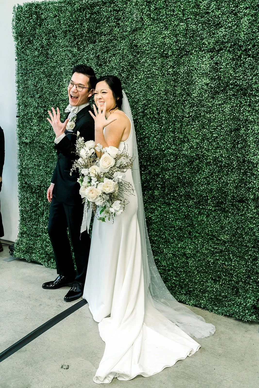 Cindy and Alex against the green hedge wall at The East Angel, her long veil trailing