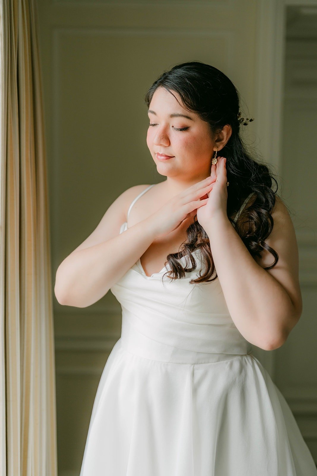Saira fastening her earring in her wedding dress by the window