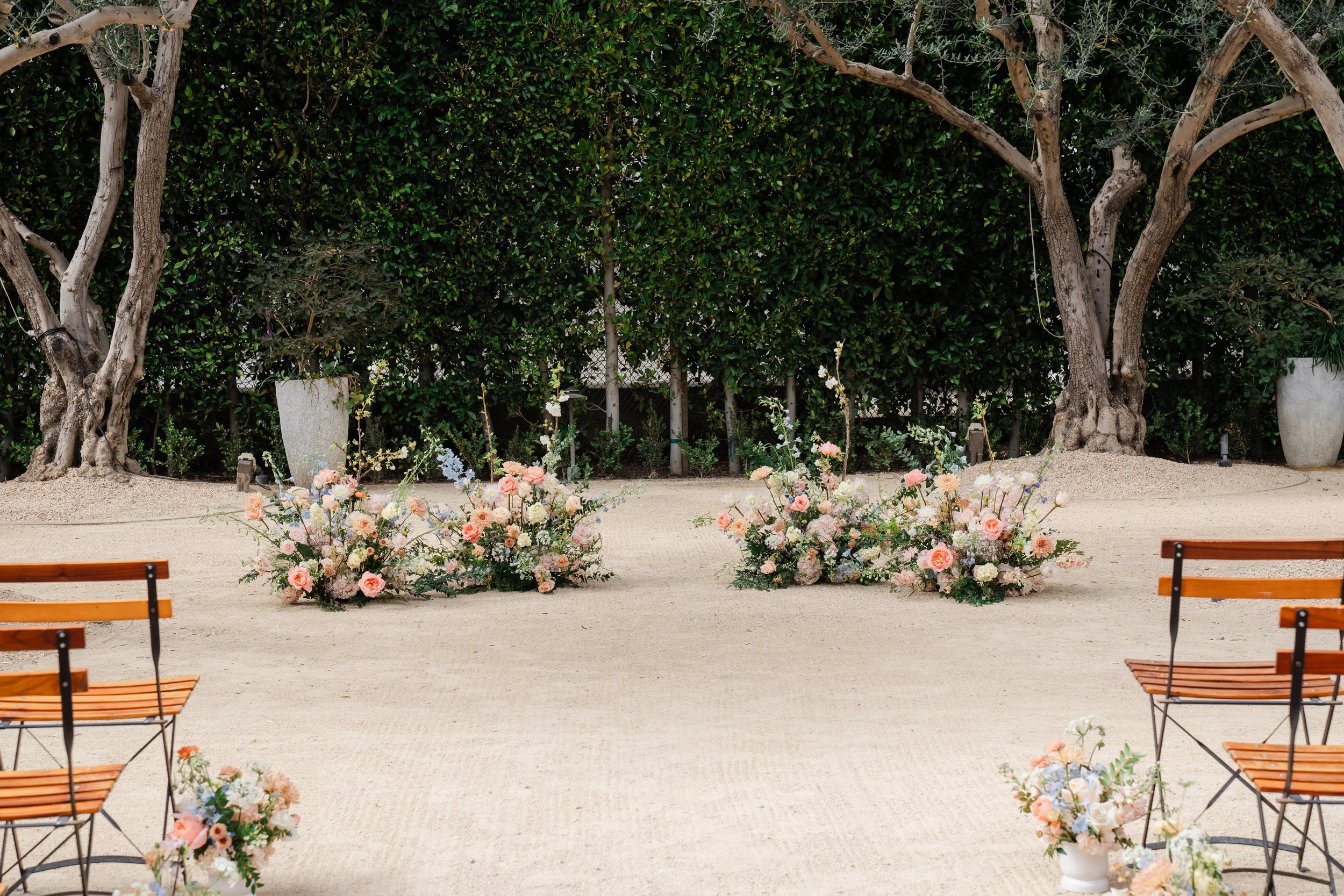 Outdoor wedding or event setup with floral arrangements, wooden benches, and large trees on a sandy ground.