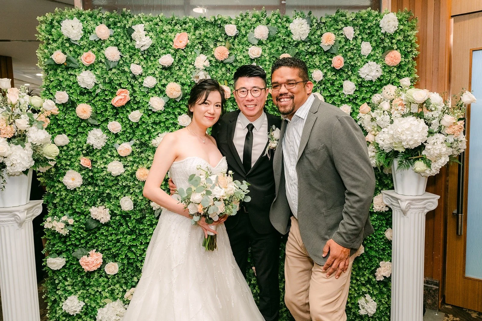 Eunice and Eric smile with a guest in front of the flower wall