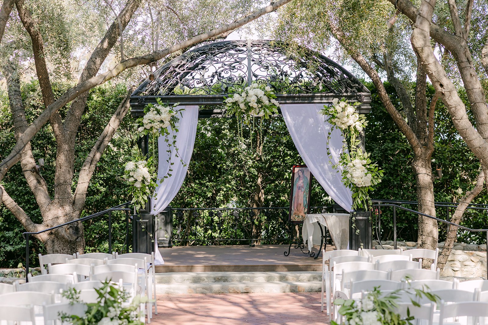 White ceremony chairs facing the gazebo beneath the olive trees