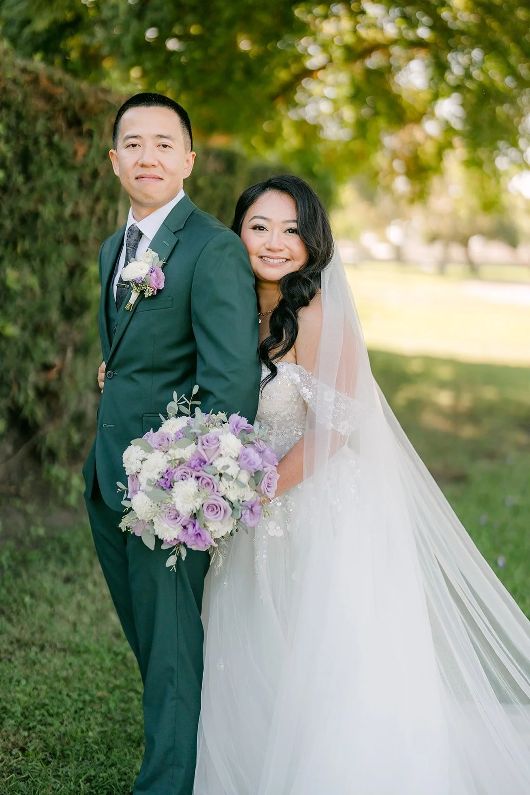 Jennifer and Kevin smile together by the hedge with her veil behind them