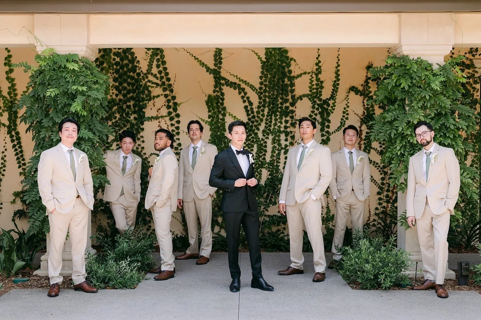 Groom in a black tux with groomsmen in tan suits against an ivy wall at Monte Bella
