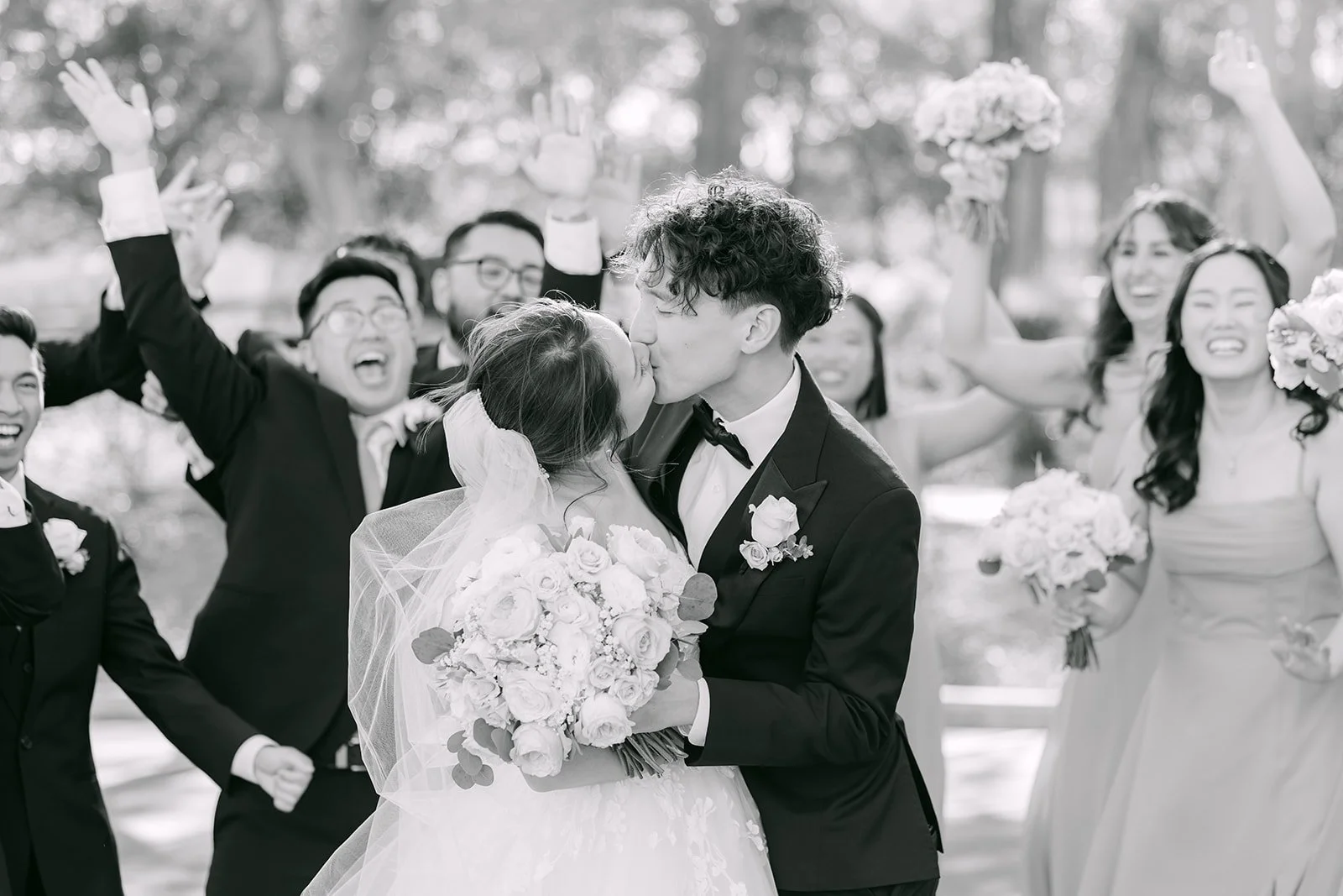 Black and white of the groom kissing the bride as the wedding party celebrates