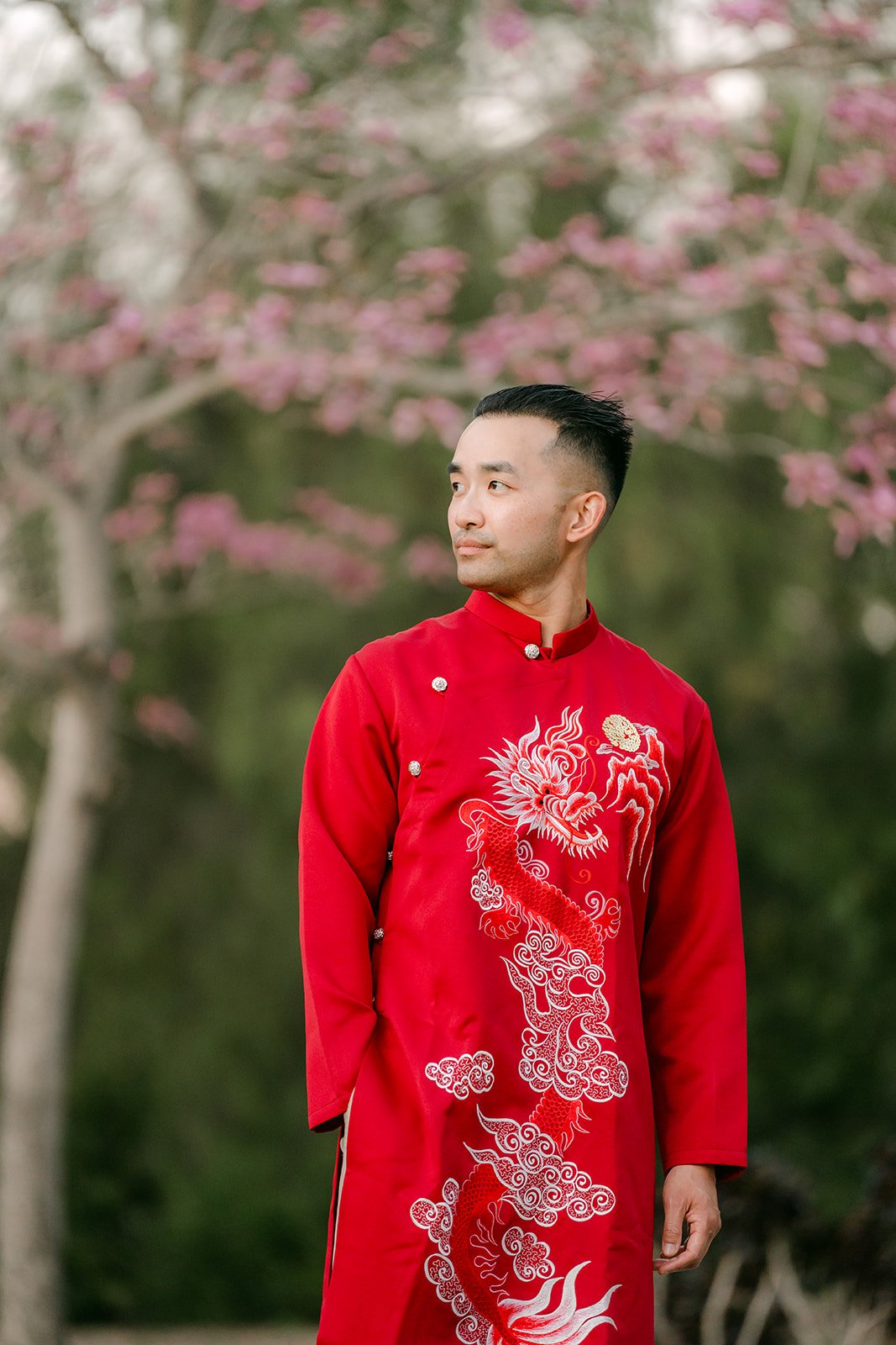 Vincent glancing to the side in his red ao dai under the flowering trees.
