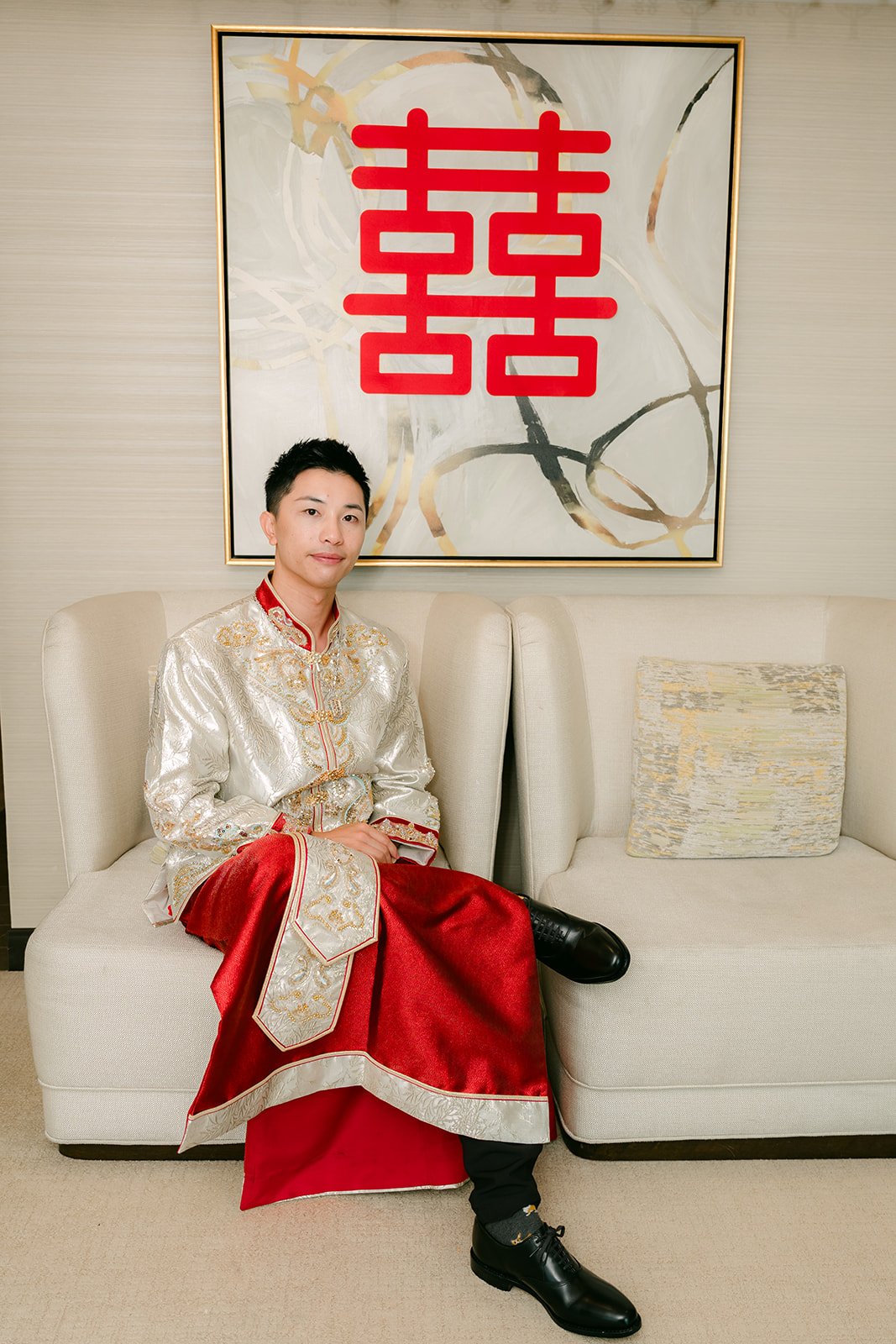 A groom seated under a double happiness painting in his traditional outfit, photographed by One and Only Production.