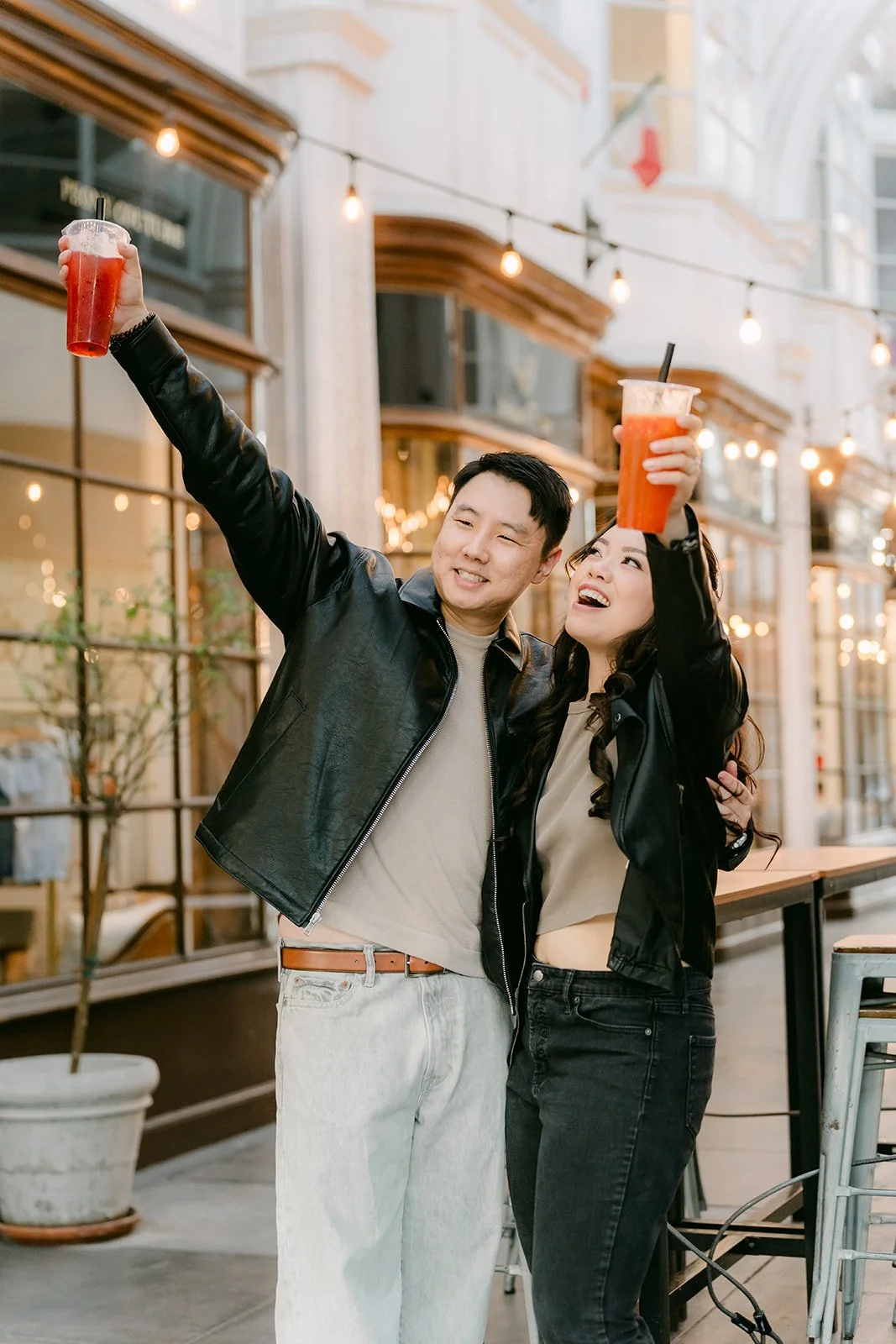 Emily and James raise their drinks in celebration beneath the string lights.