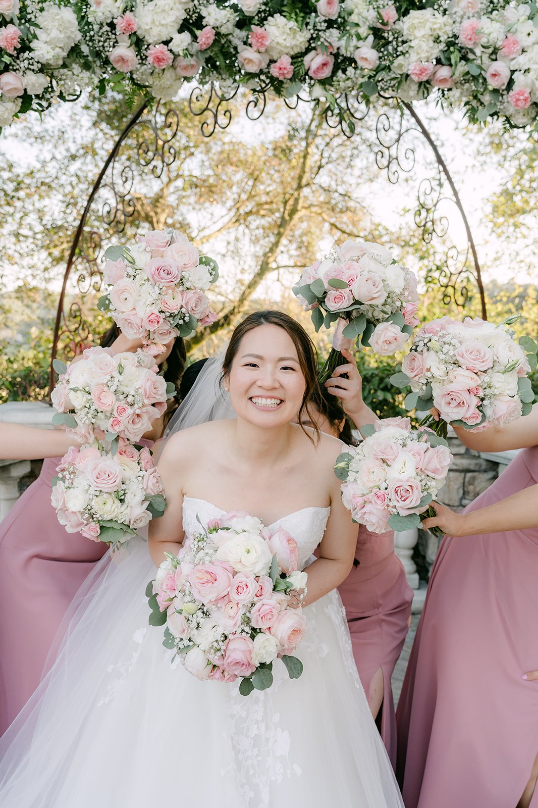 Bride surrounded by bridesmaids' bouquets under the floral arch