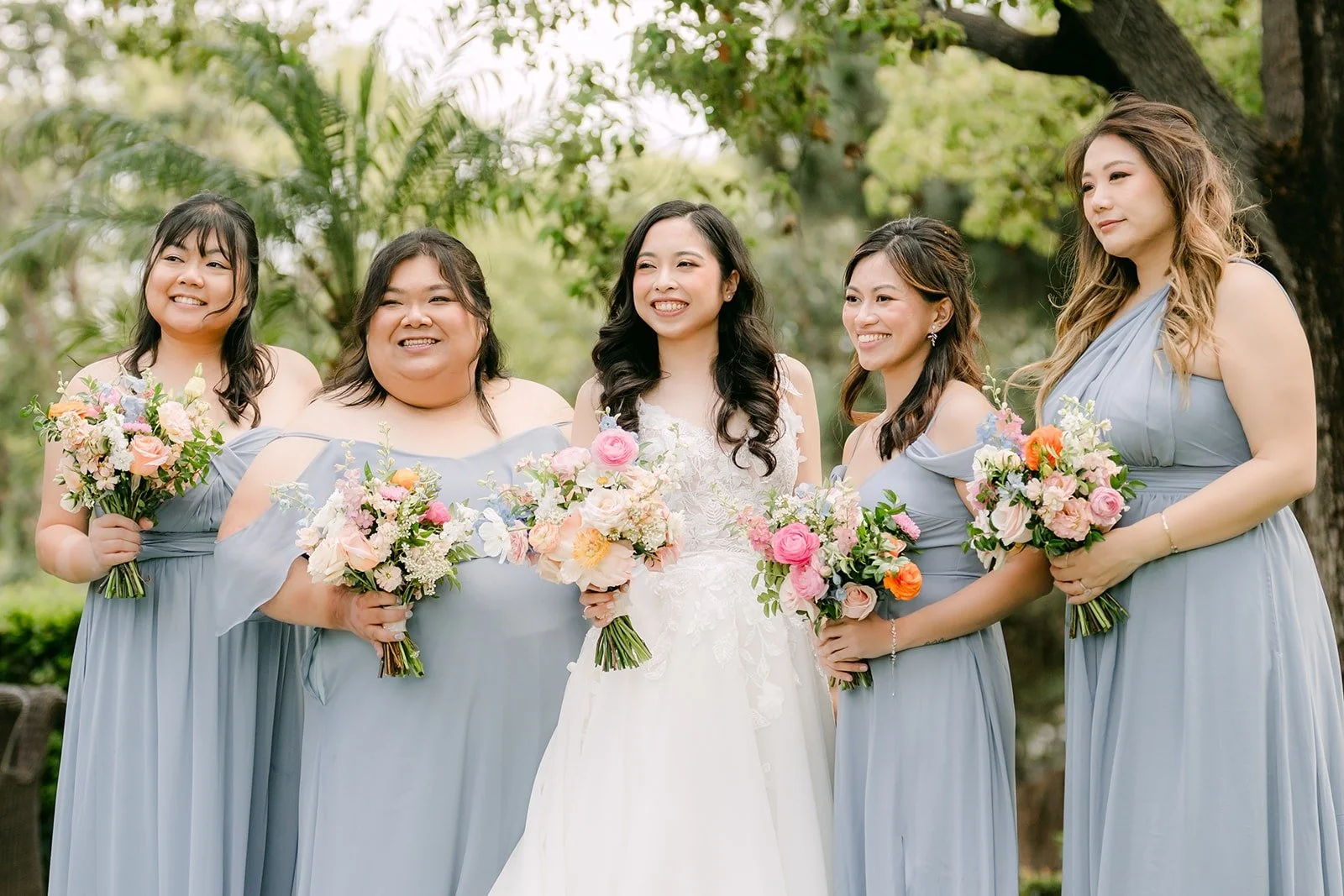Natalie with her bridesmaids in dusty blue holding pastel bouquets under the trees