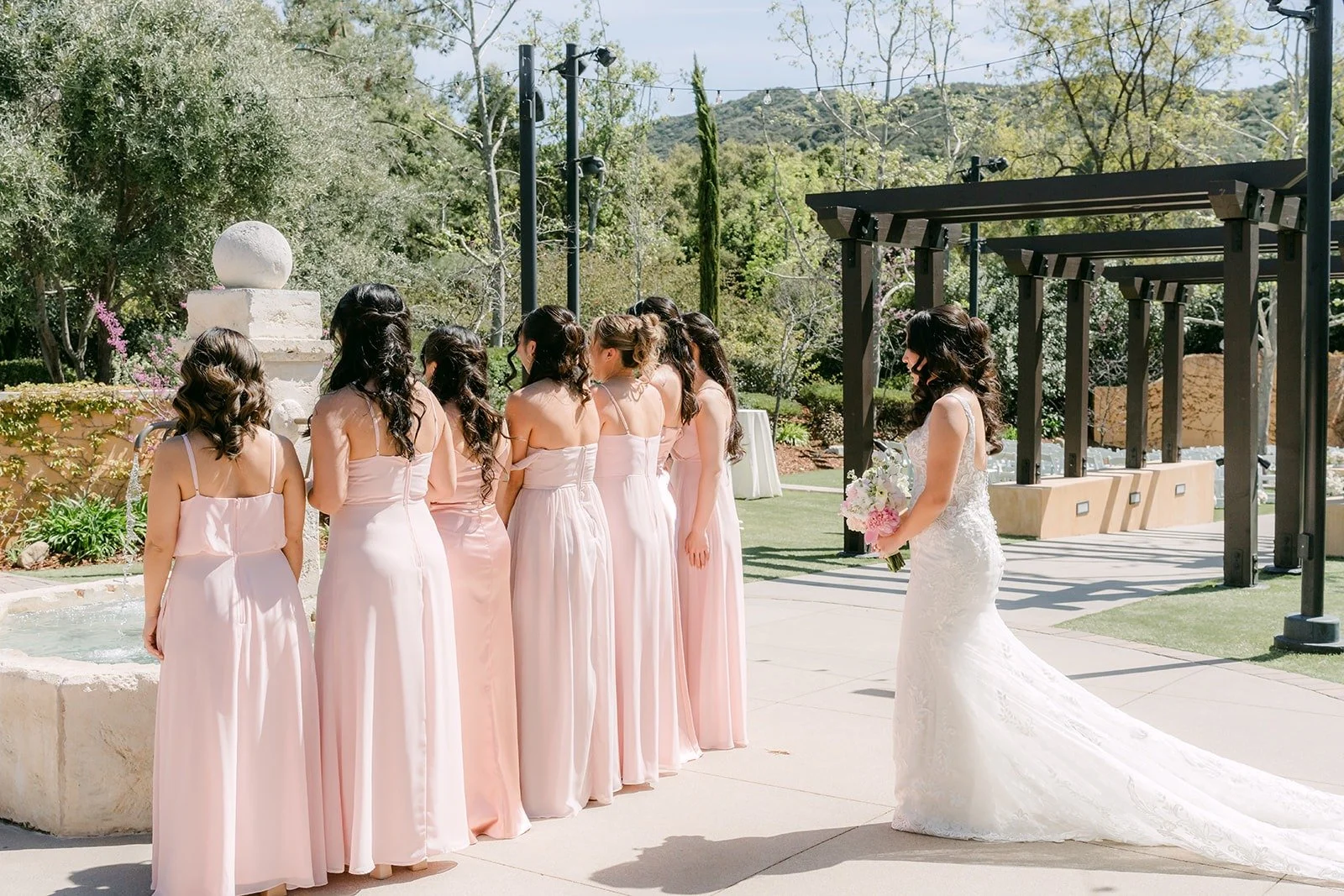 Kelli's bridesmaids in blush dresses seeing her in her gown for the first time by the pergola