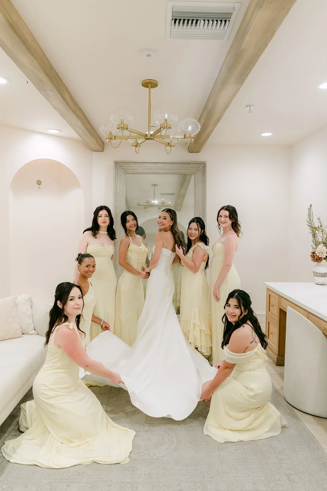 Bride and bridesmaids in butter yellow arranging her gown under the chandelier