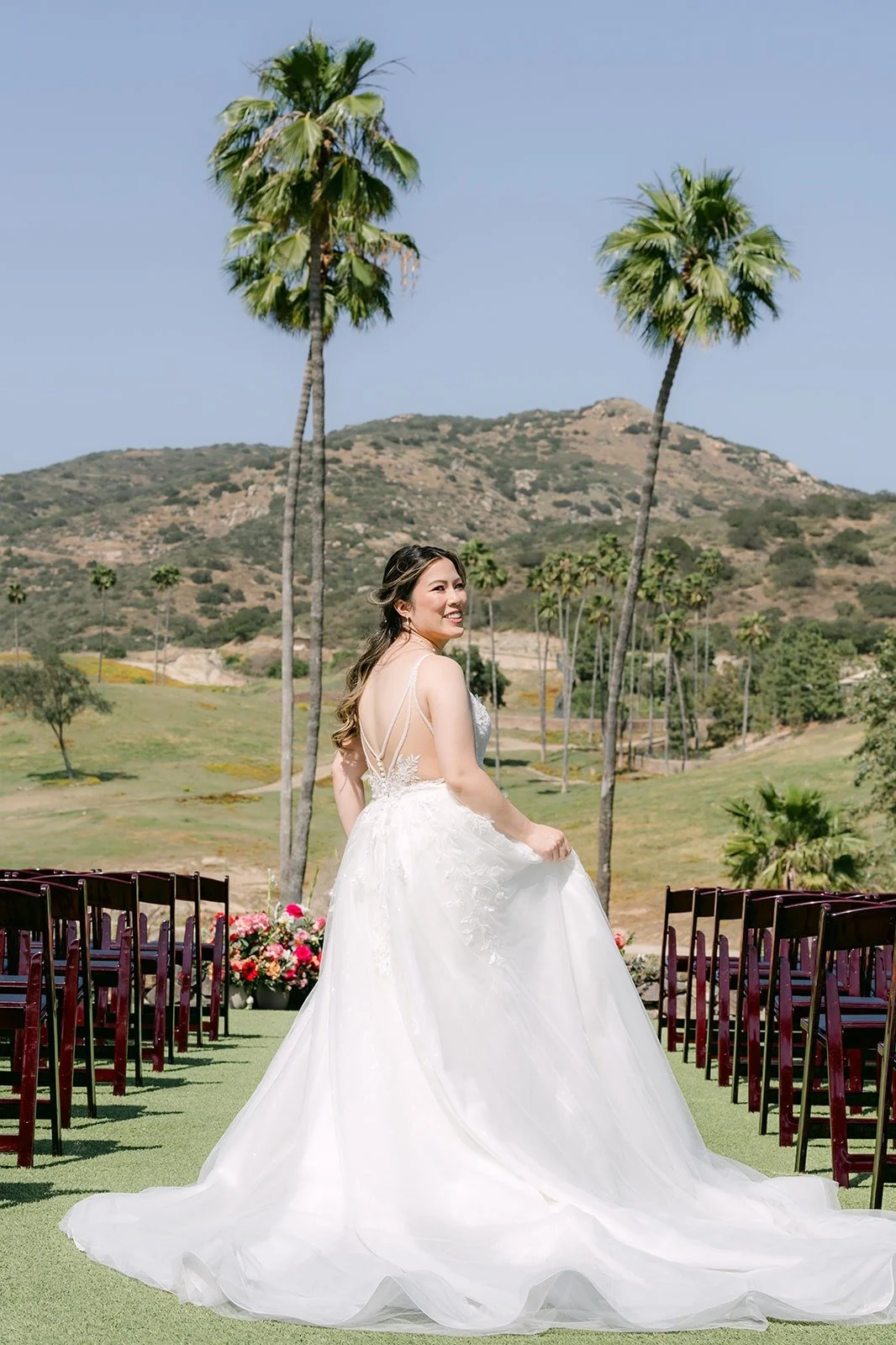 Joanne in her gown and long train on the ceremony lawn with palm trees and hills behind her