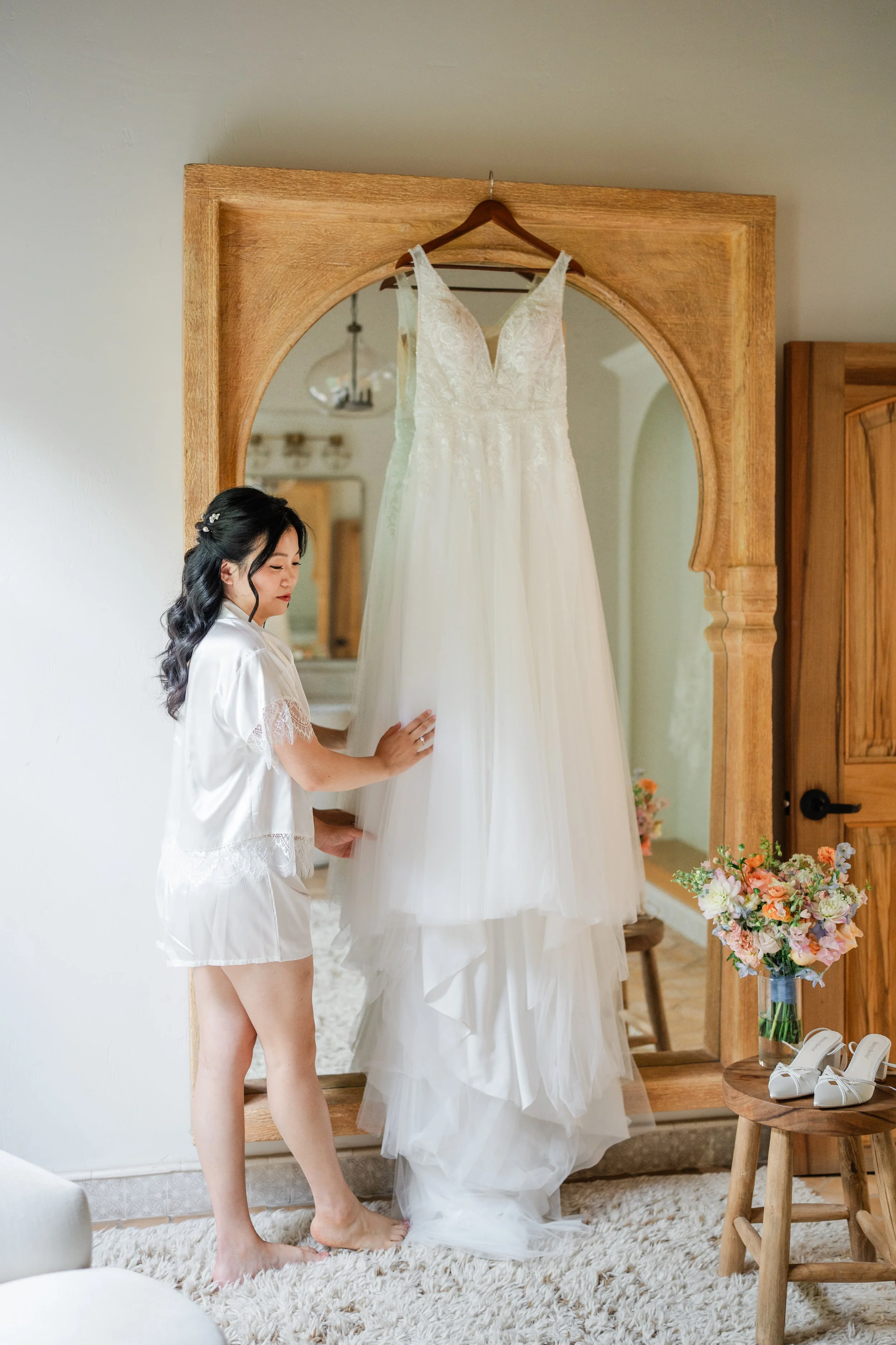 A woman in a satin robe stands in front of a large mirror, touching a hanging white wedding dress with lace details, in a warmly lit room decorated with flowers.