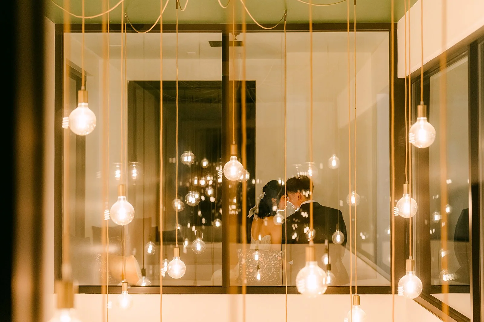Vivian and Alex framed by a window and hanging lights at night