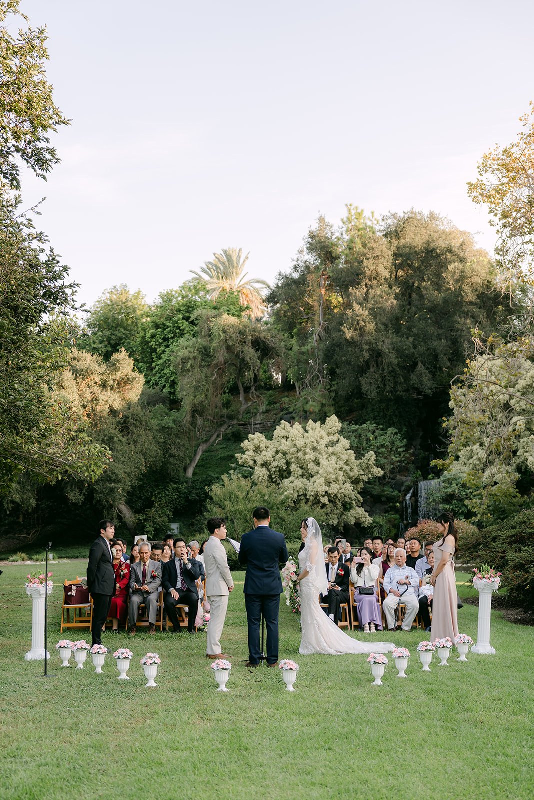 Wide view of the ceremony on the lawn with trees and pedestal florals.