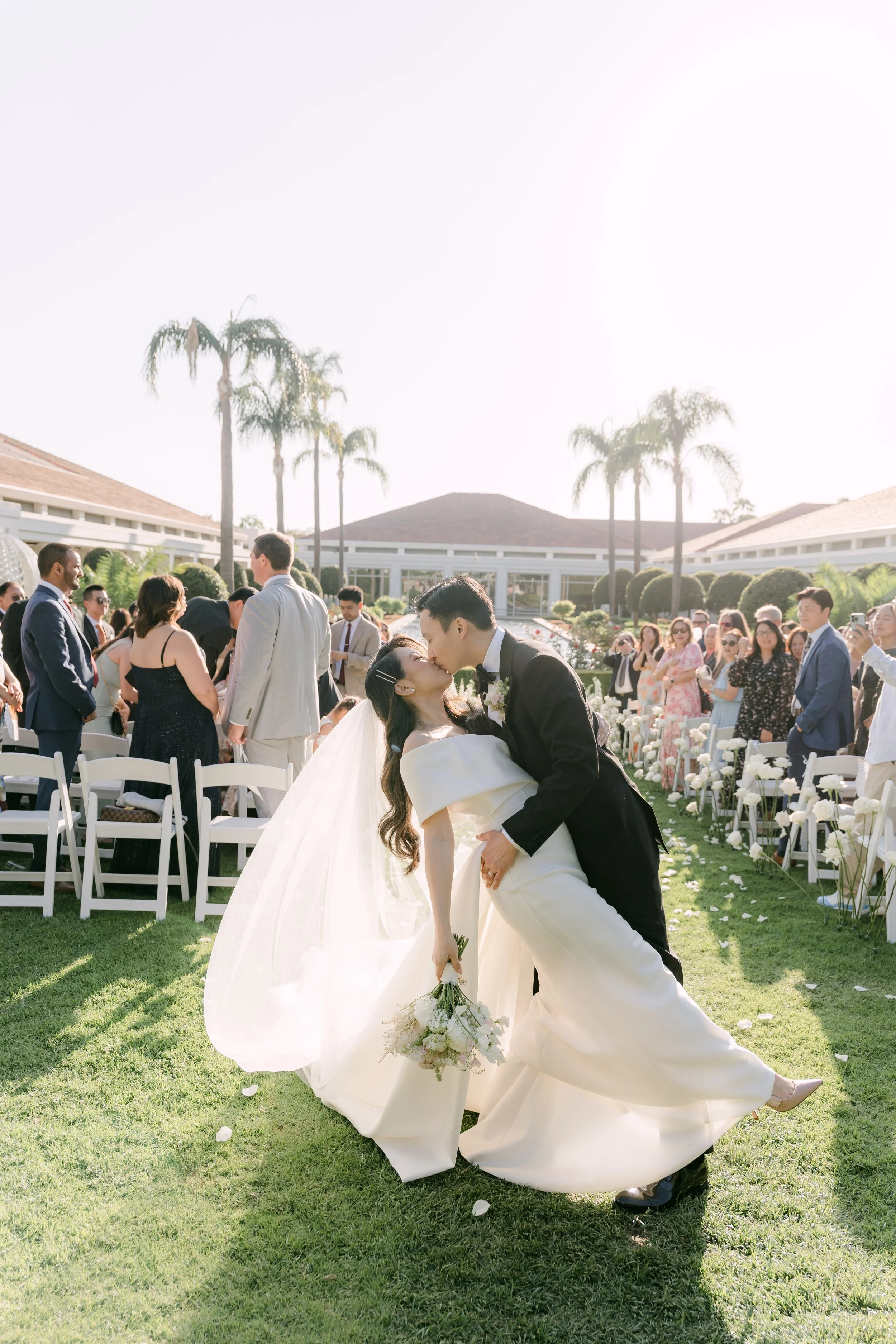A newlywed couple sharing a kiss during their outdoor wedding ceremony, with guests watching and smiling in the background. The scene is sunny, with palm trees, chairs, white flowers, and a modern building in the background.