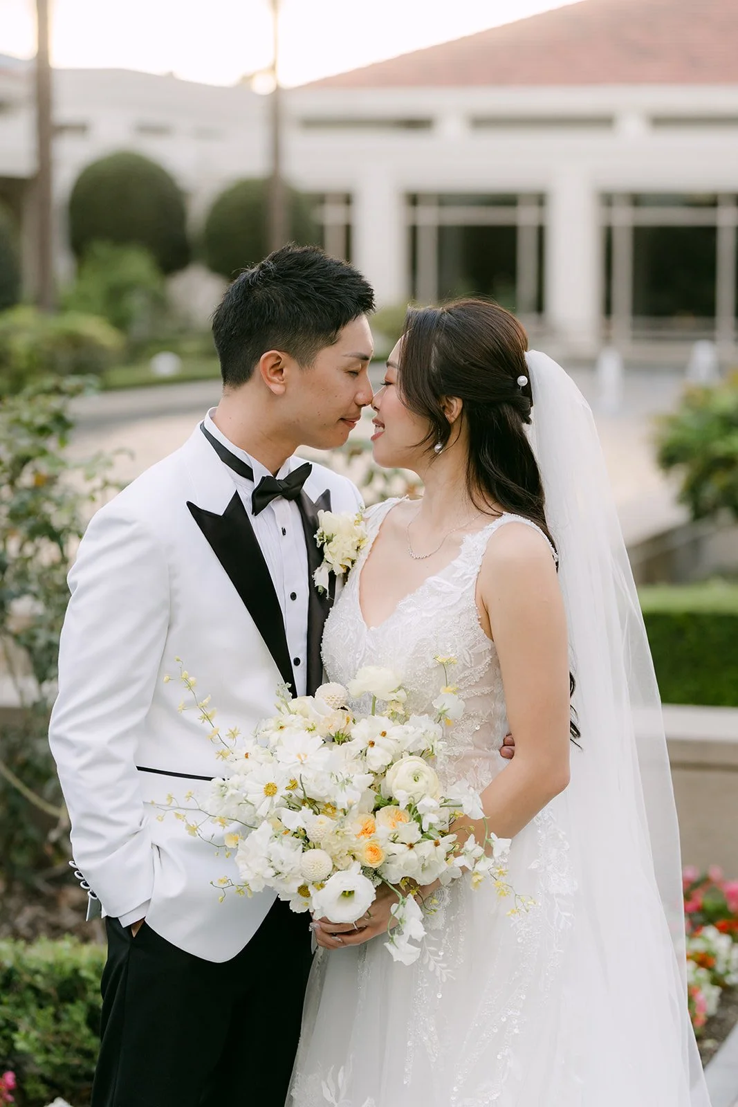 Jenny and Ryan kissing with her bouquet in front of a white building and palms.