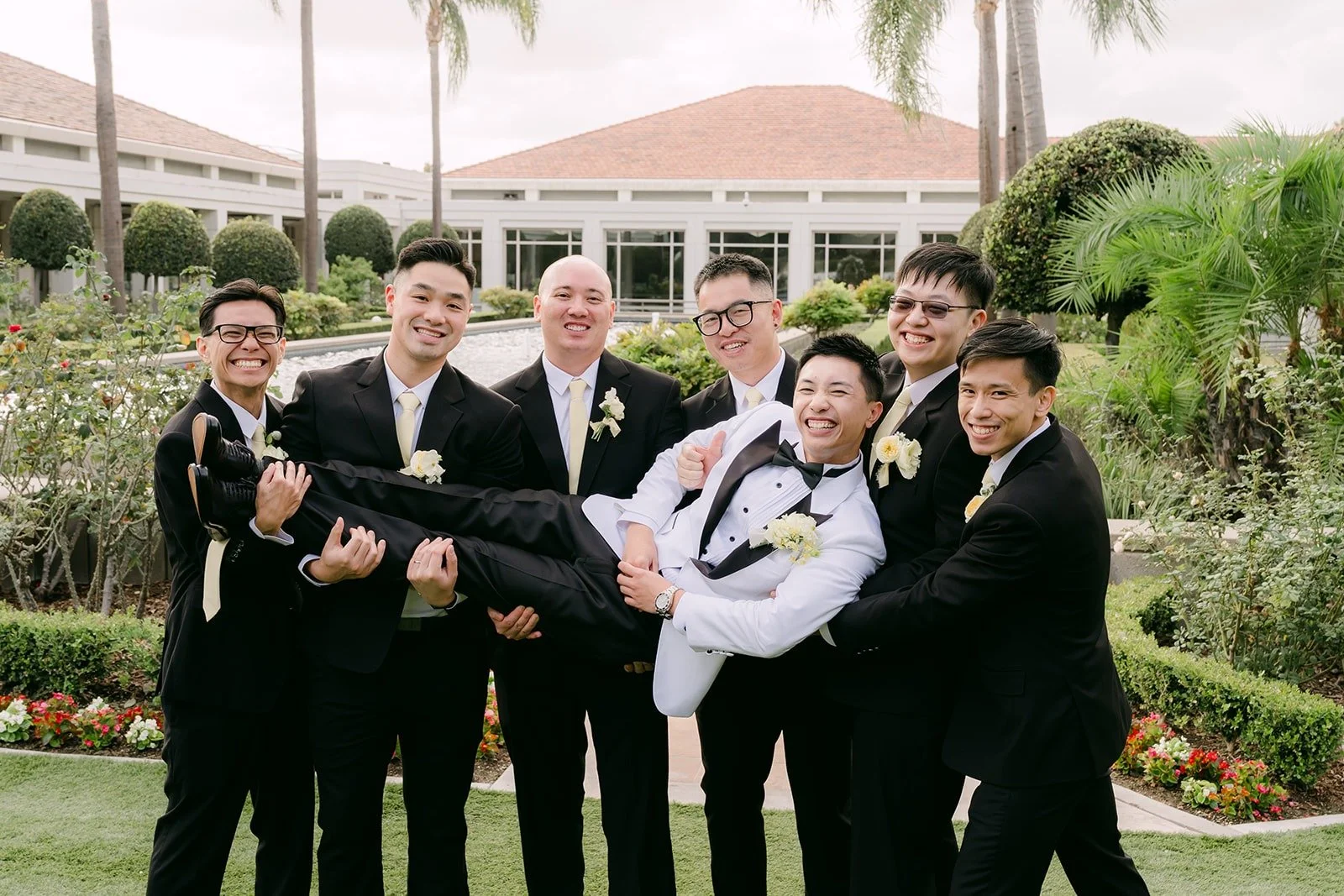 Groomsmen holding Ryan up sideways in front of the palm trees, photographed by One and Only Production.