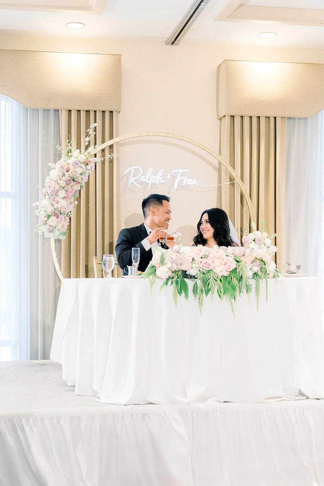 Francesca and Ralph at their sweetheart table under a floral hoop