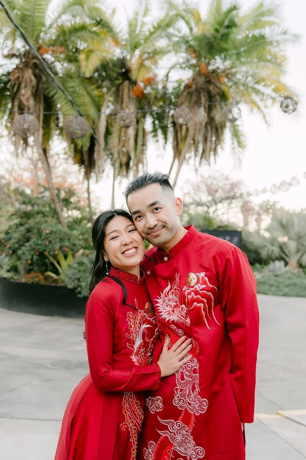 Kim and Vincent in matching red ao dai smiling cheek to cheek under the palms, photographed by One and Only Production.