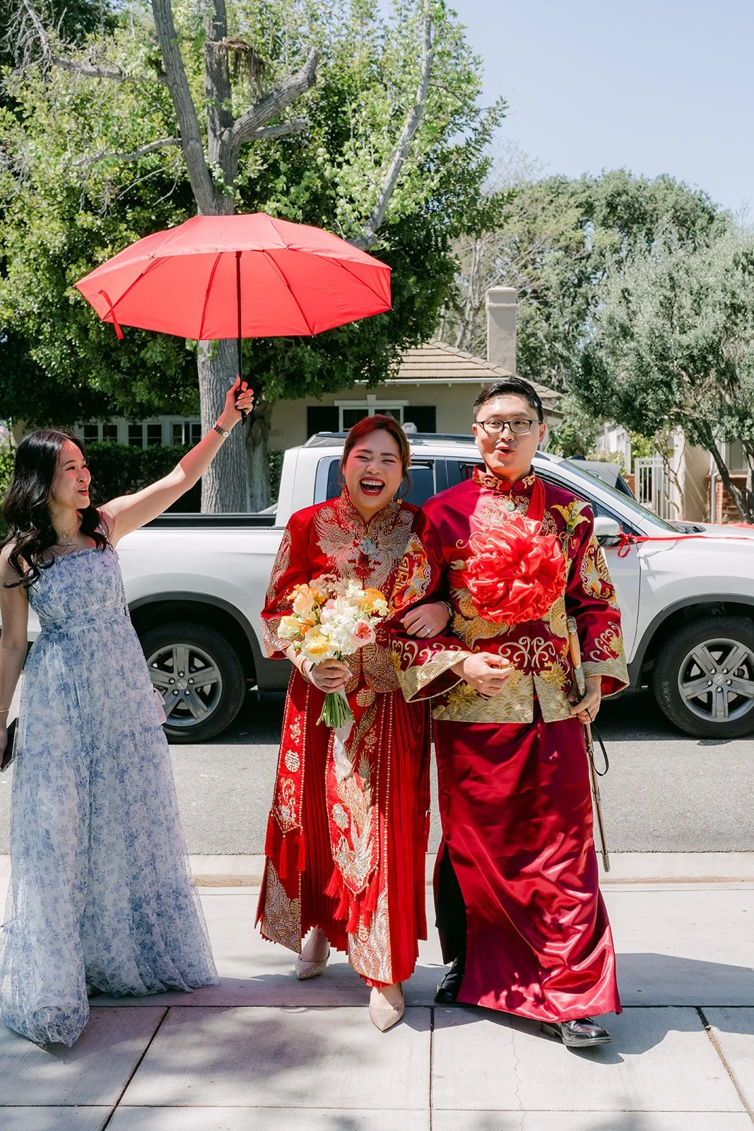 The bride and groom walking under a red umbrella in front of the house.