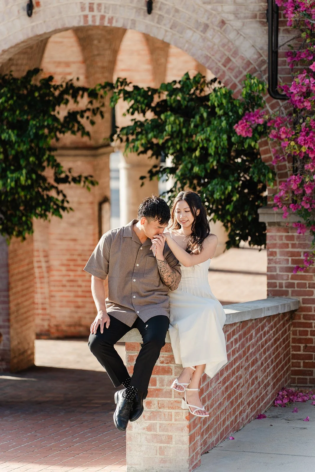 Andrew sits on a brick wall with Victoria under the archway and bougainvillea.