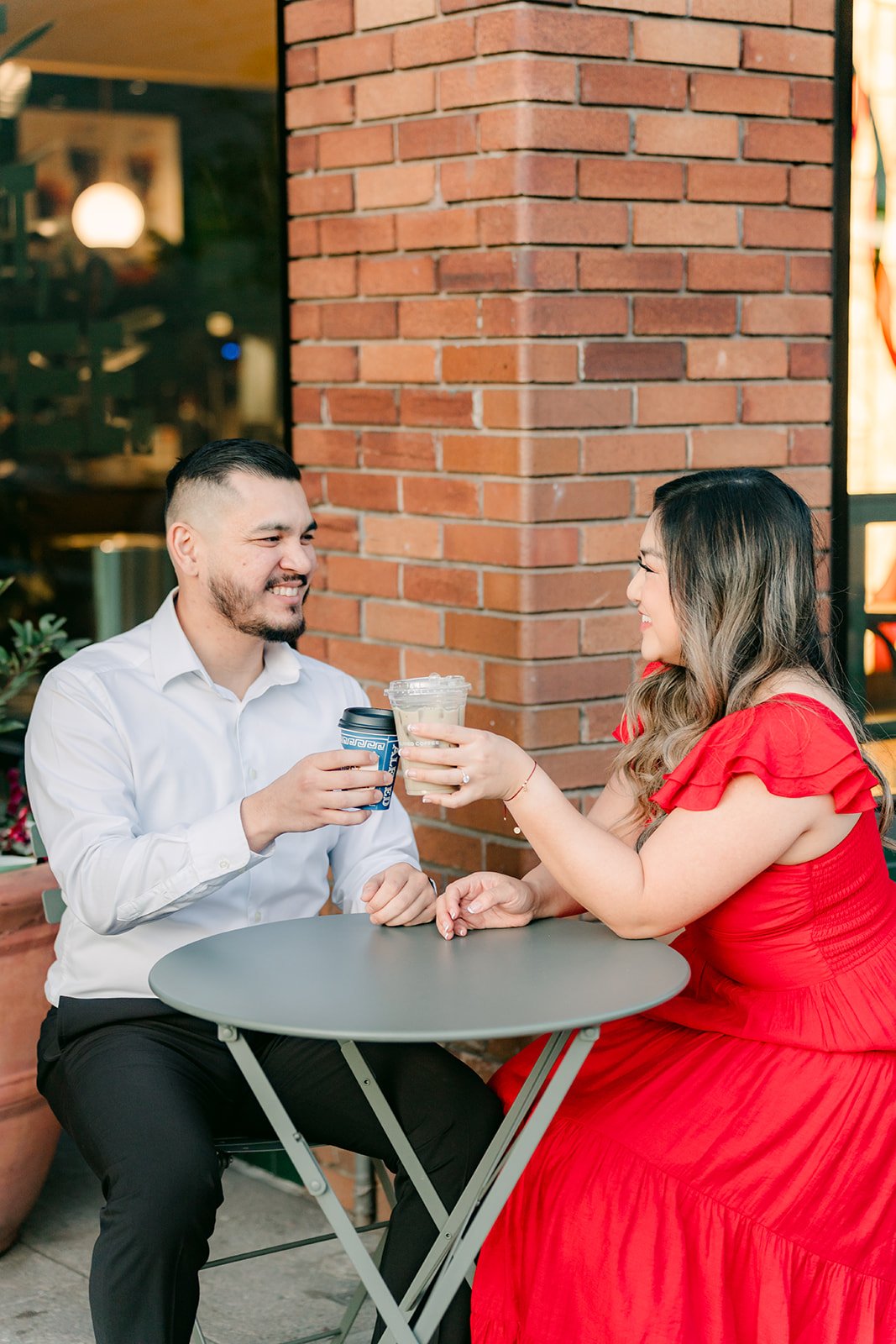 Linh and Ankush toast with coffee cups at a sidewalk cafe table.