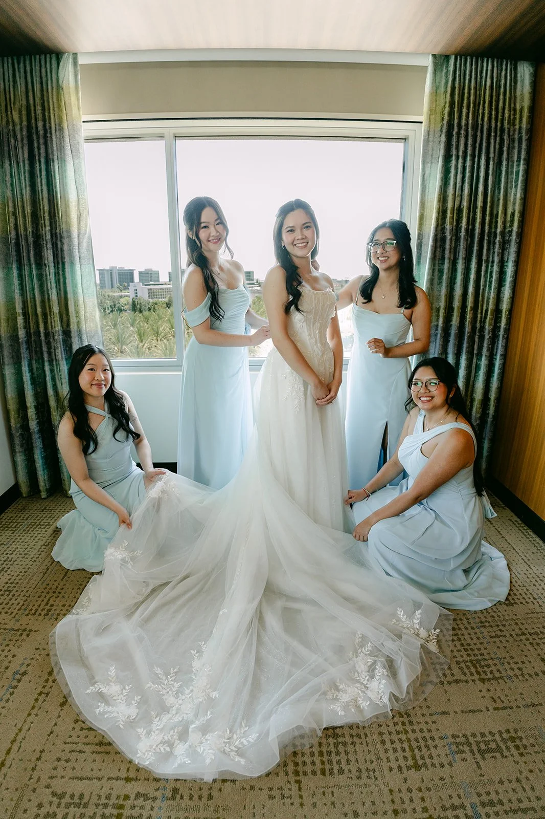 Bridesmaids in light blue dresses kneeling around the bride's train in the bridal suite