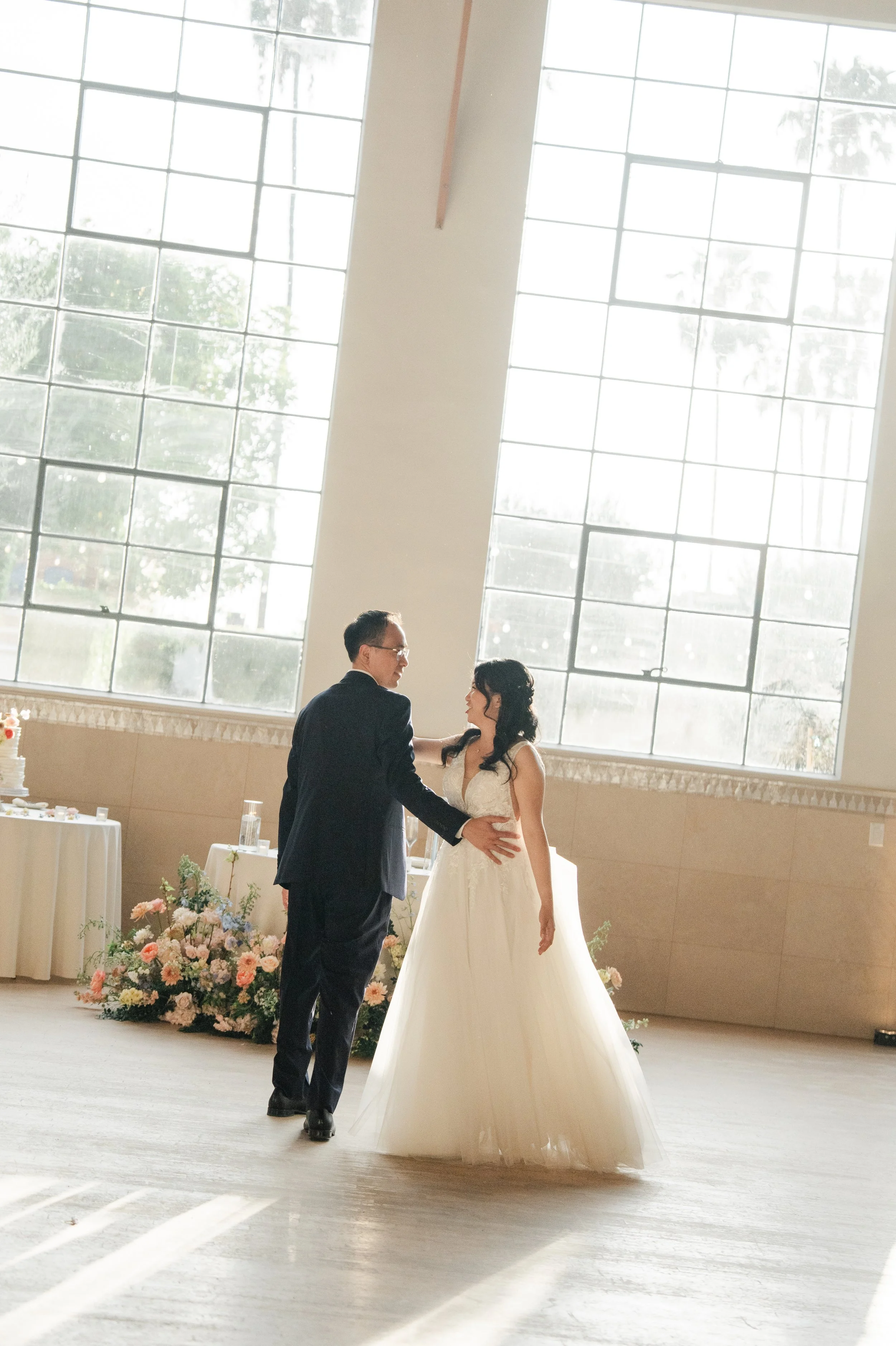 A bride and groom are dancing in a wedding reception hall with large windows, decorated with flowers and a wedding cake in the background.