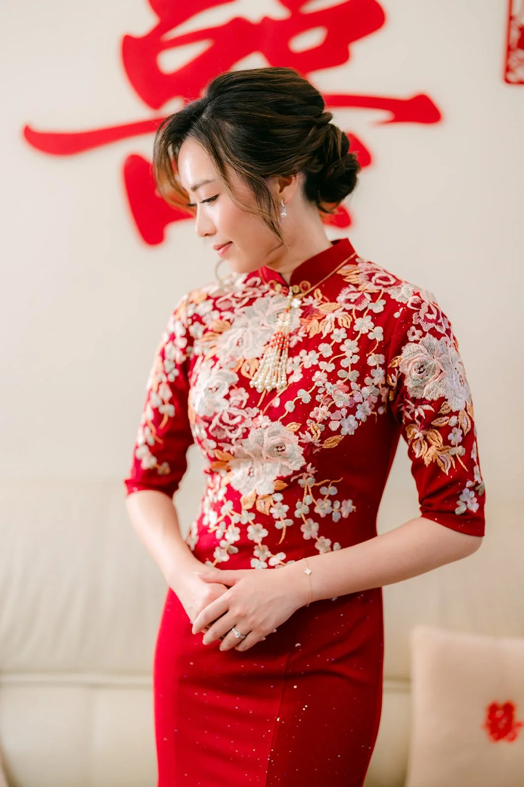 A bride smiling in her red qipao with a double happiness sign behind her.