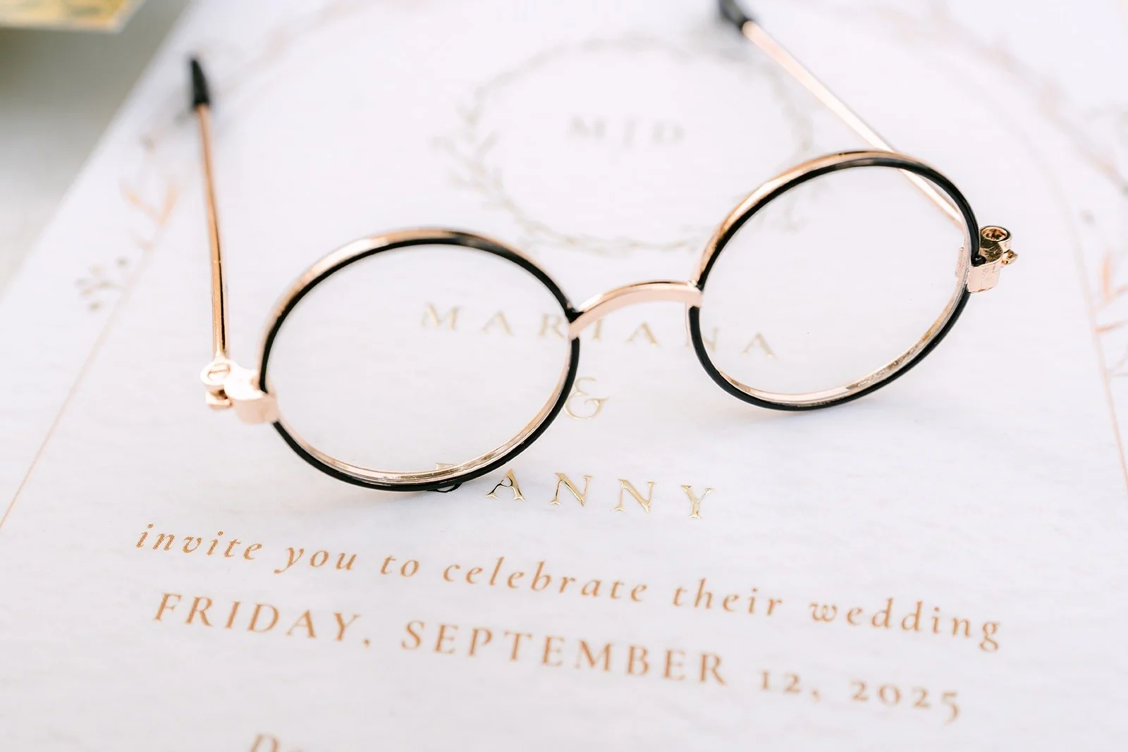 Round gold glasses resting on Mariana and Danny's invitation for Friday, September 12, 2025