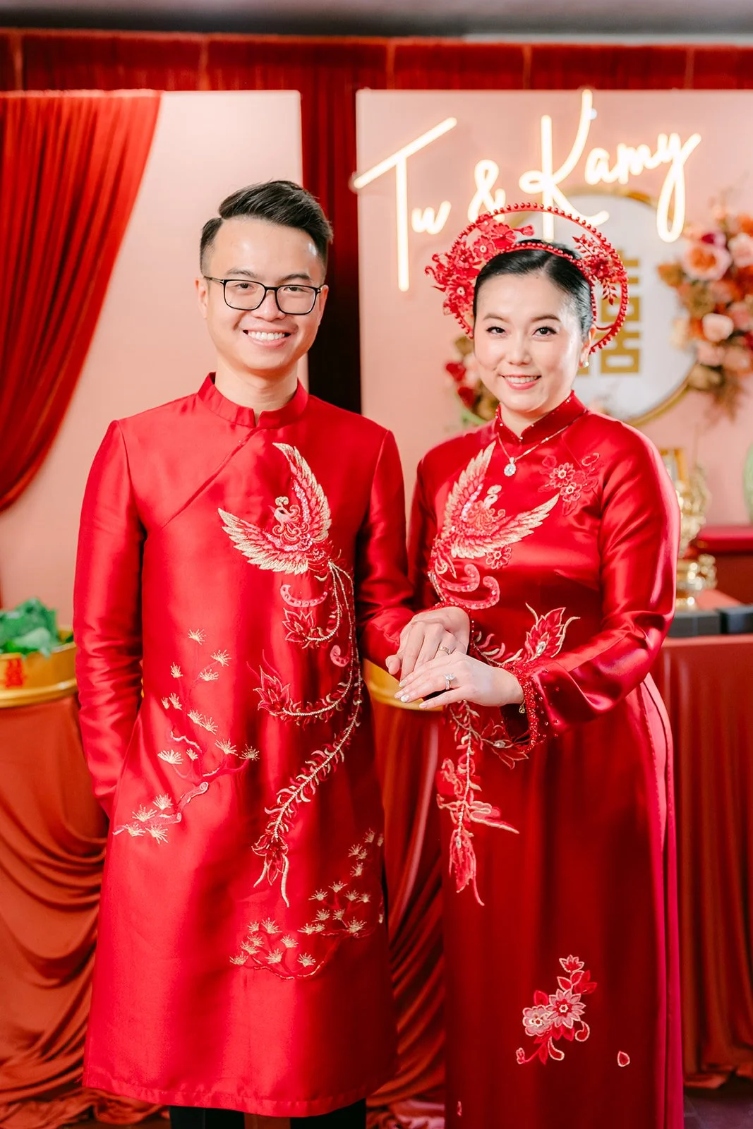 A couple in matching red ao dai standing in front of their tea ceremony backdrop.