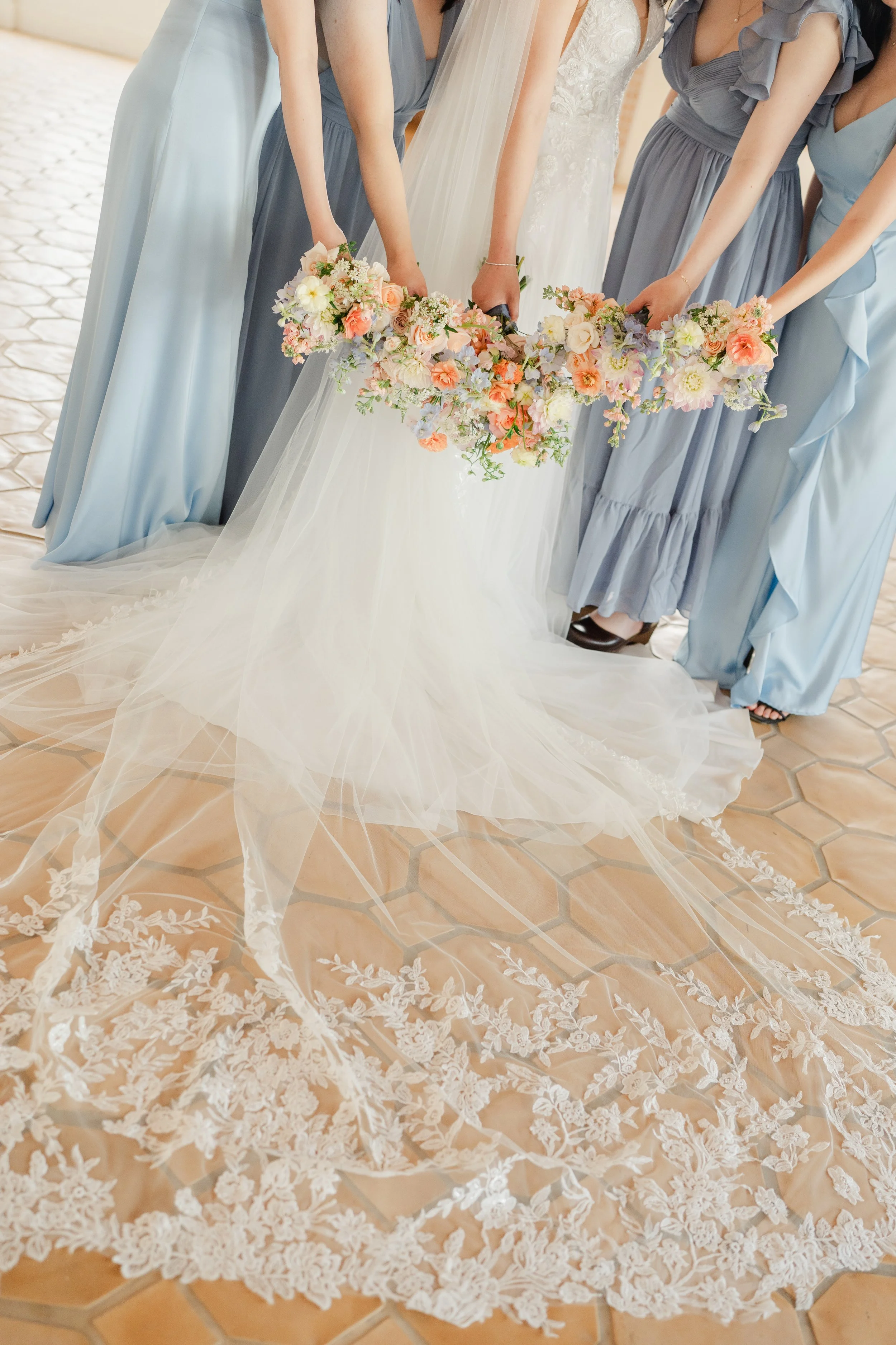 A group of women, including a bride in a white wedding dress and bridesmaids in blue dresses, holding a floral wedding bouquet with pink, white, and cream flowers, standing on a tiled floor.
