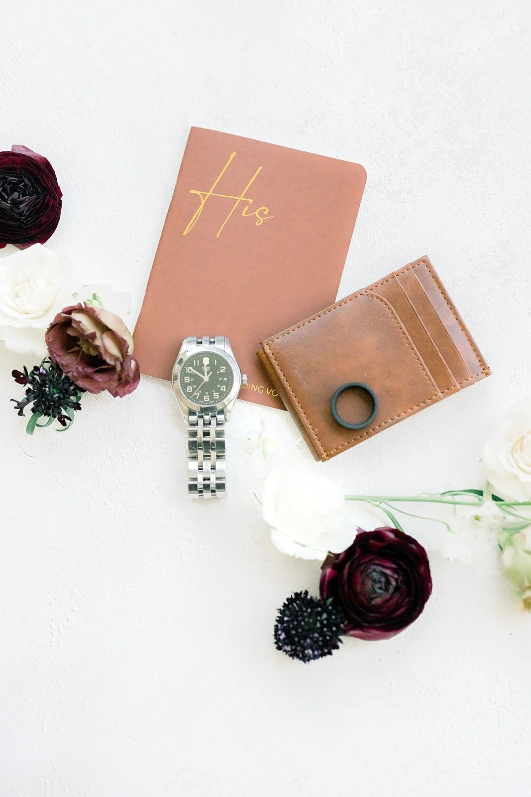 A tan leather vow book with Mikey's watch and rings among burgundy flowers