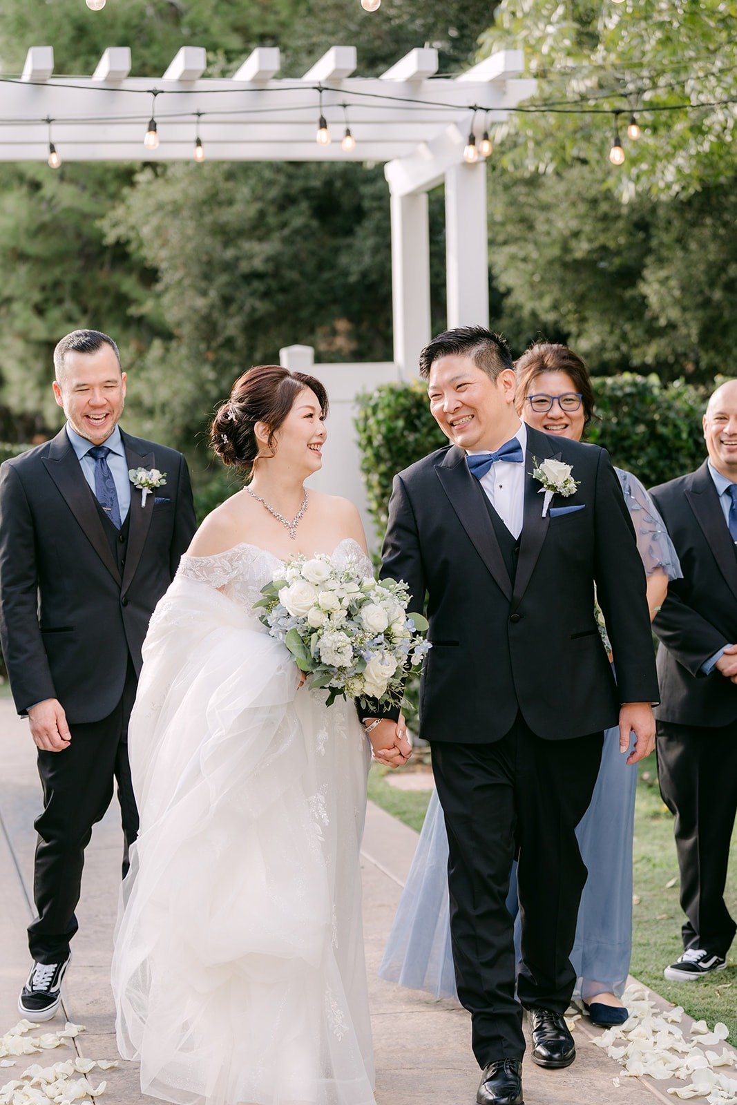 Sochi and Jay walk with their wedding party past the white arbor