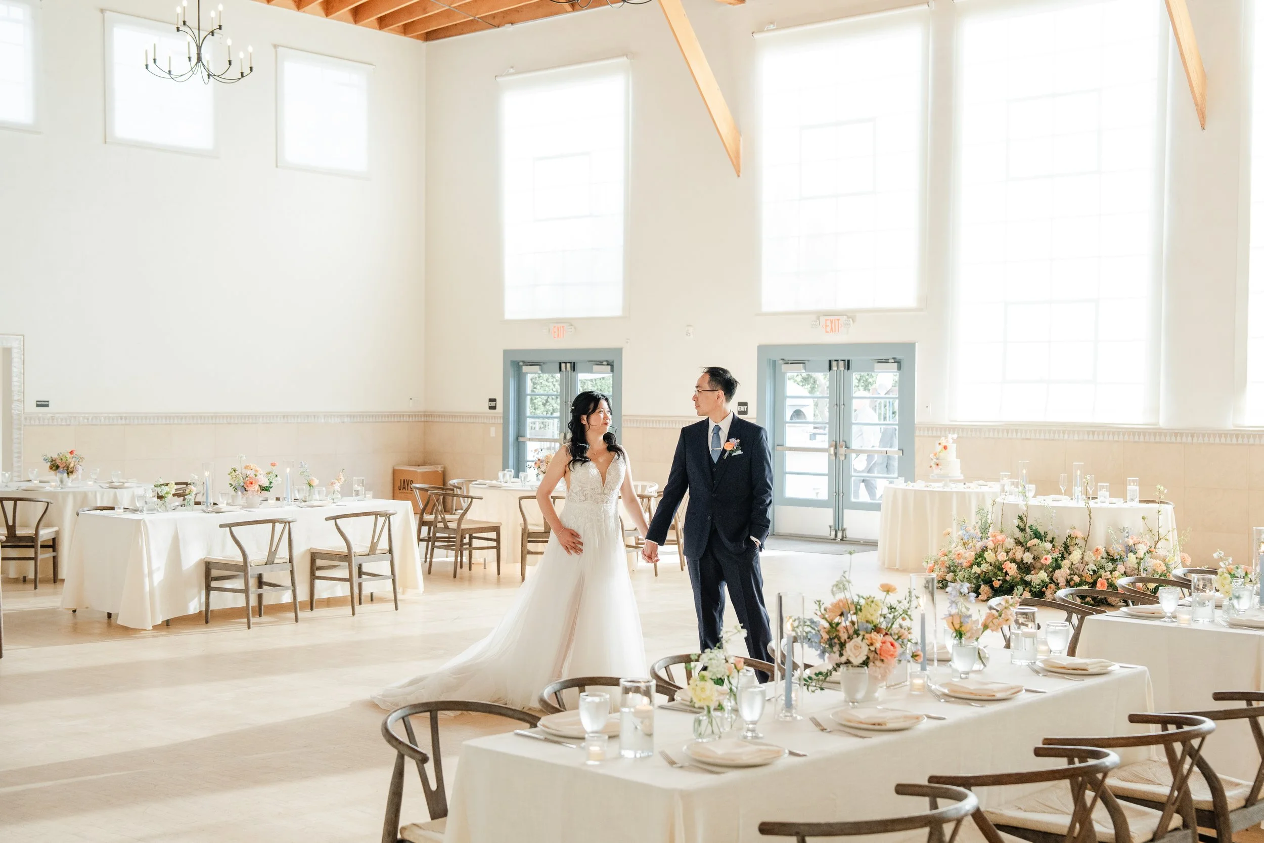 A bride and groom walk hand in hand inside a bright, airy wedding reception hall decorated with floral centerpieces and round tables with white tablecloths.