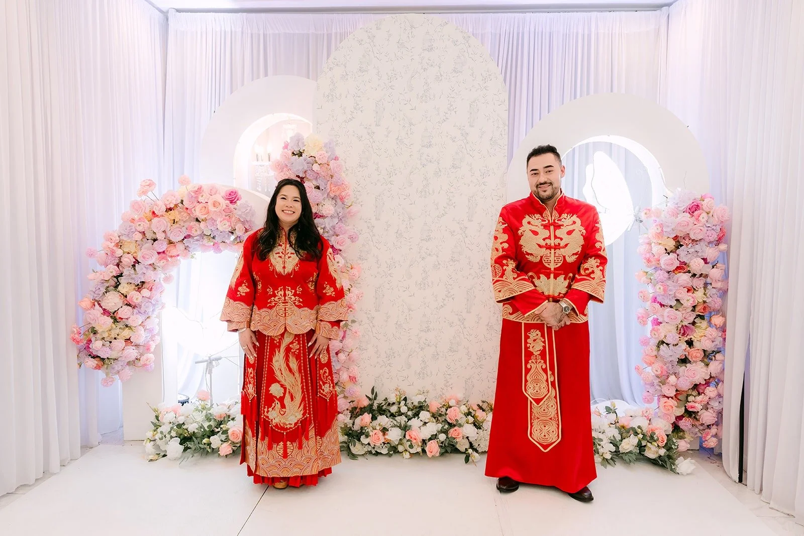 Amy and James in red embroidered outfits in front of the floral arch