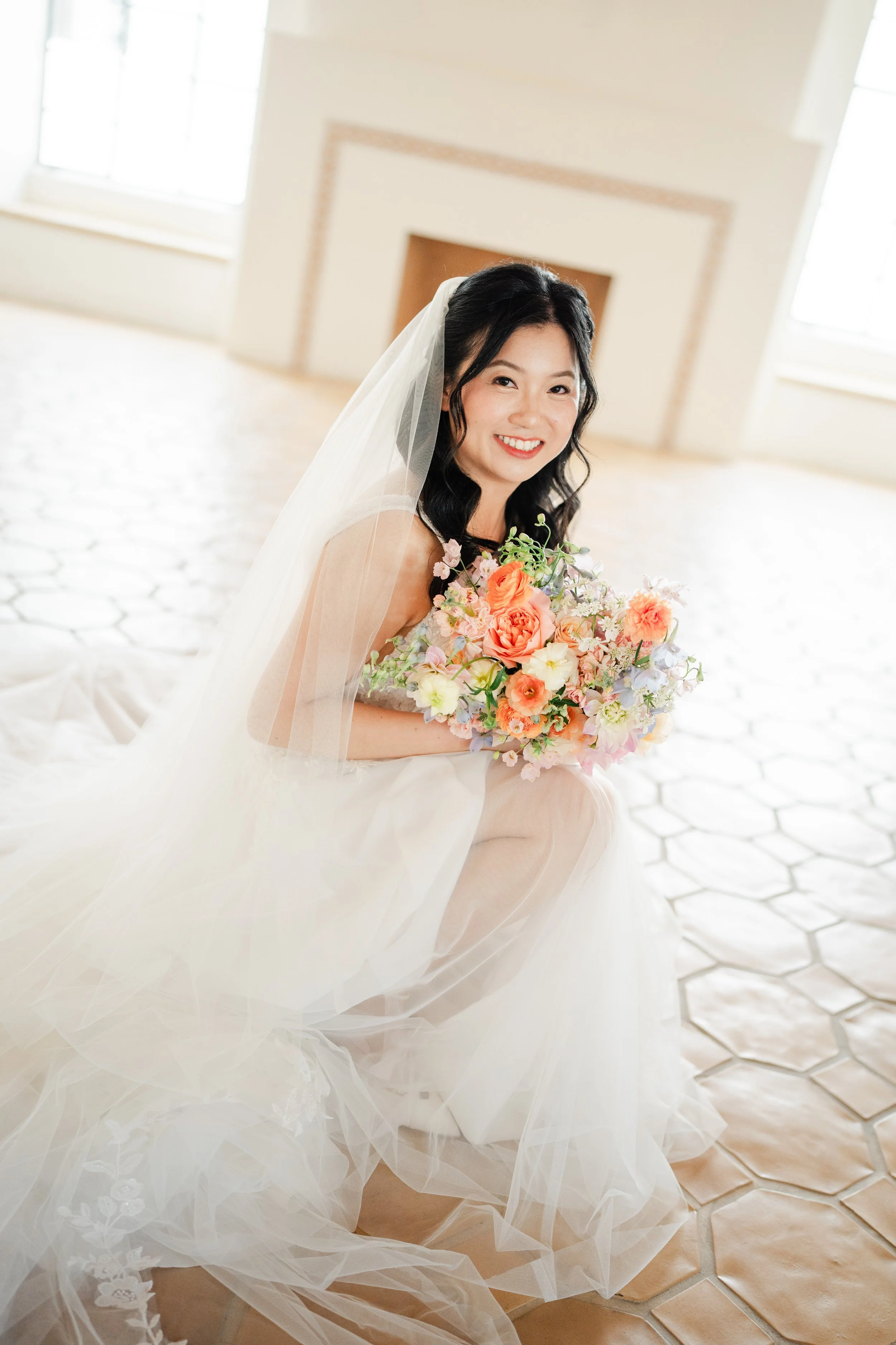 A smiling Asian bride with black hair in a strapless white wedding gown holding a bouquet of pink, white, and peach flowers inside a bright room with large windows and a tiled floor.