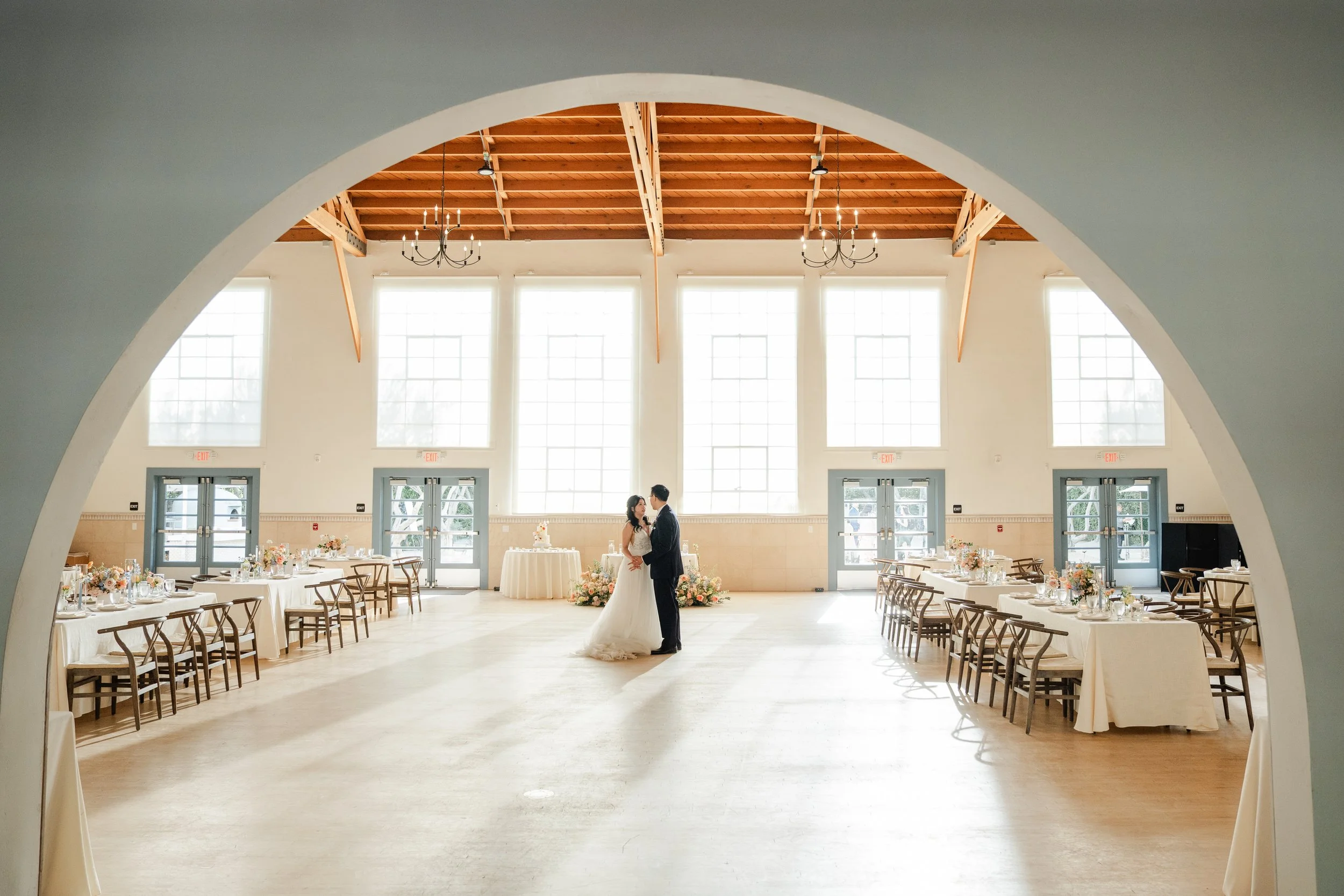 A bride and groom in wedding attire standing close together in a spacious, well-lit reception hall with high windows and wooden ceiling beams.