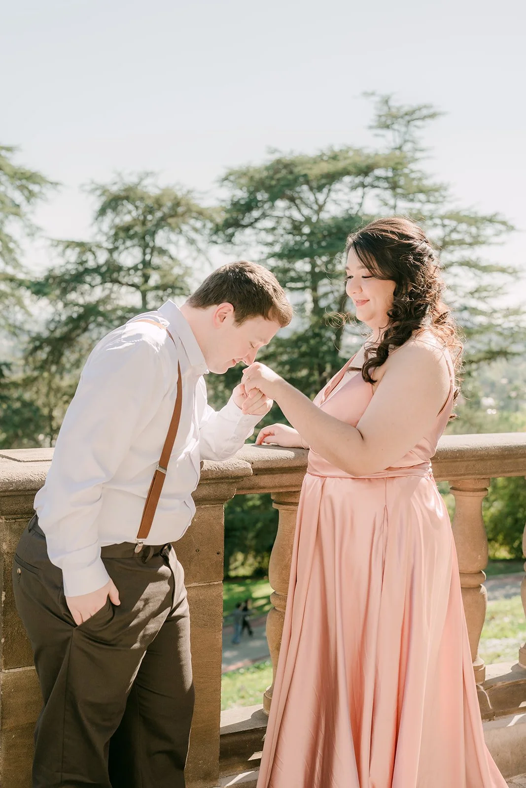 Matthew kisses Saira's hand at the stone balustrade with pines behind them.