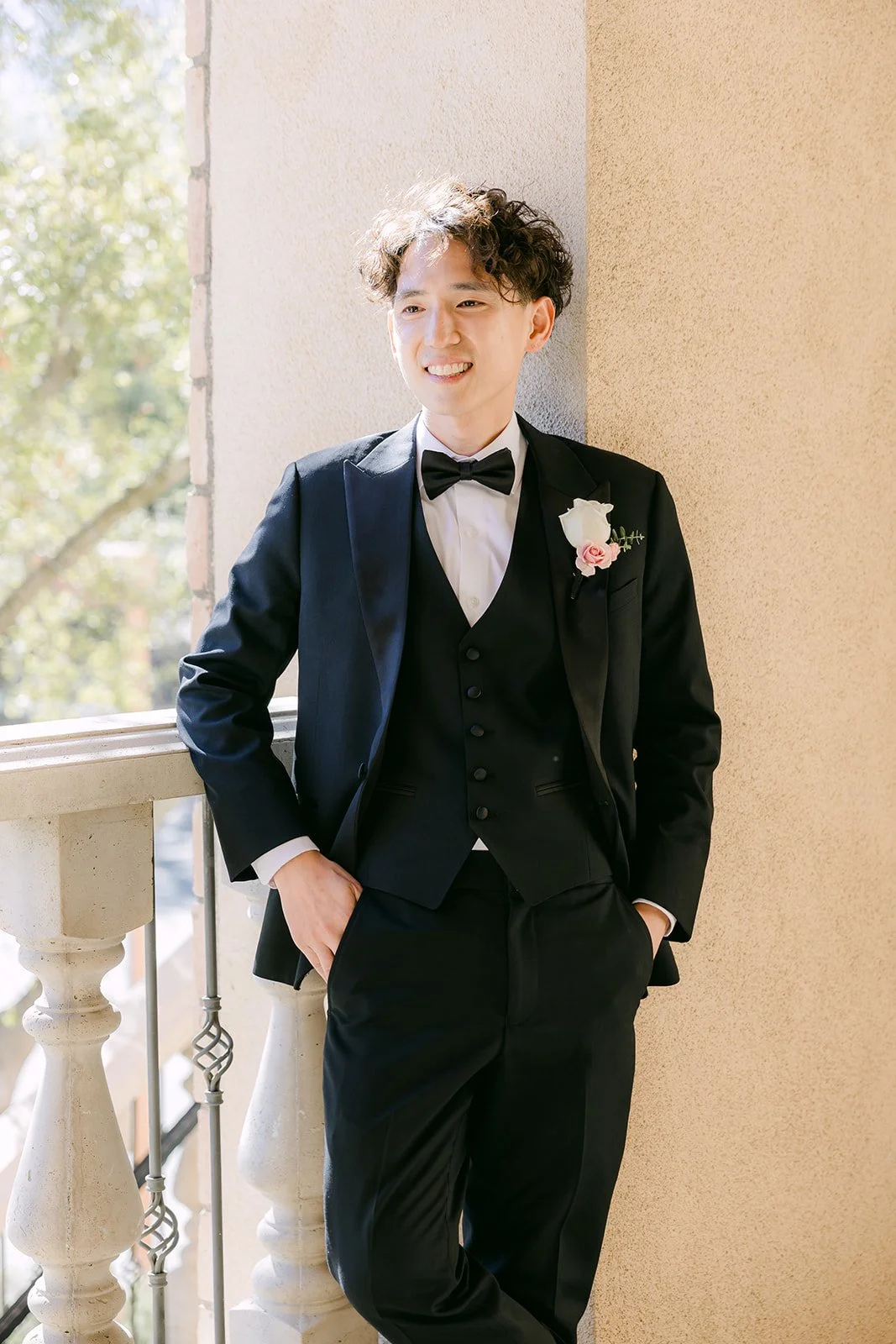 Groom leaning against a sunny balcony column in his tuxedo