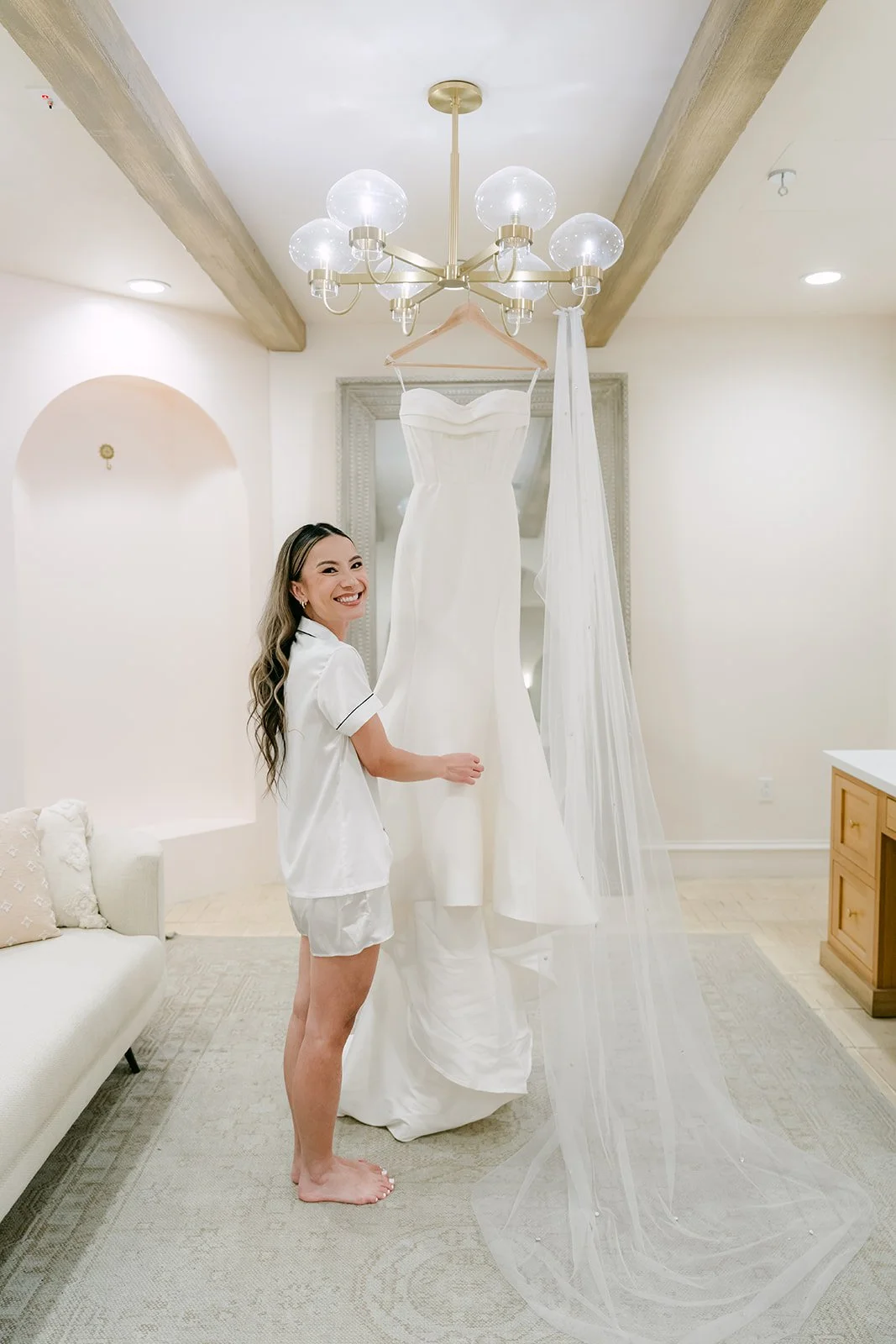 Bride in a white robe smiling beside her wedding dress in the Serra Plaza bridal suite
