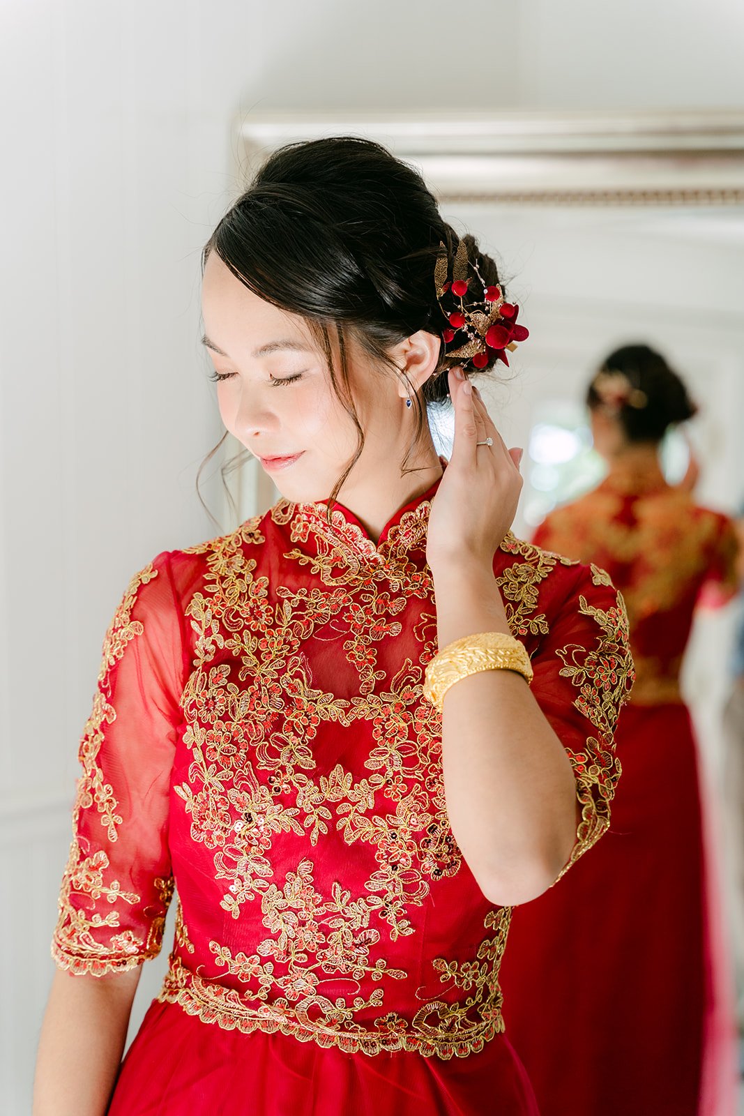 A bride in a red and gold qun kwa adjusting her hair piece by the window.